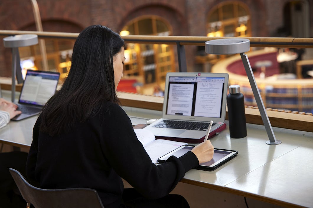 En person sitter vid ett skrivbord i ett bibliotek med en laptop, surfplatta, anteckningsbok och vattenflaska.