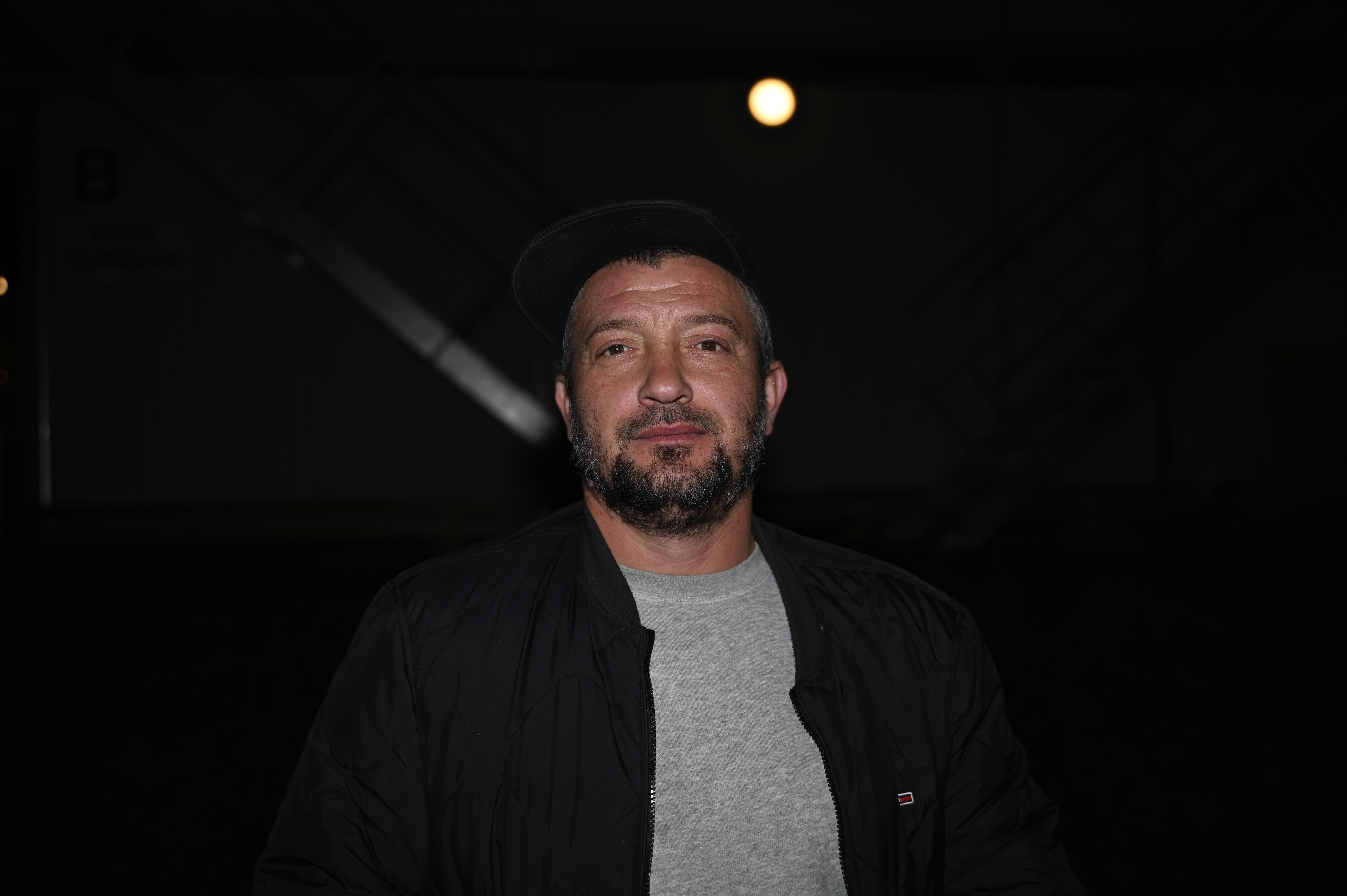 En man med keps och jacka står utomhus på natten med en ljuskälla i bakgrunden.