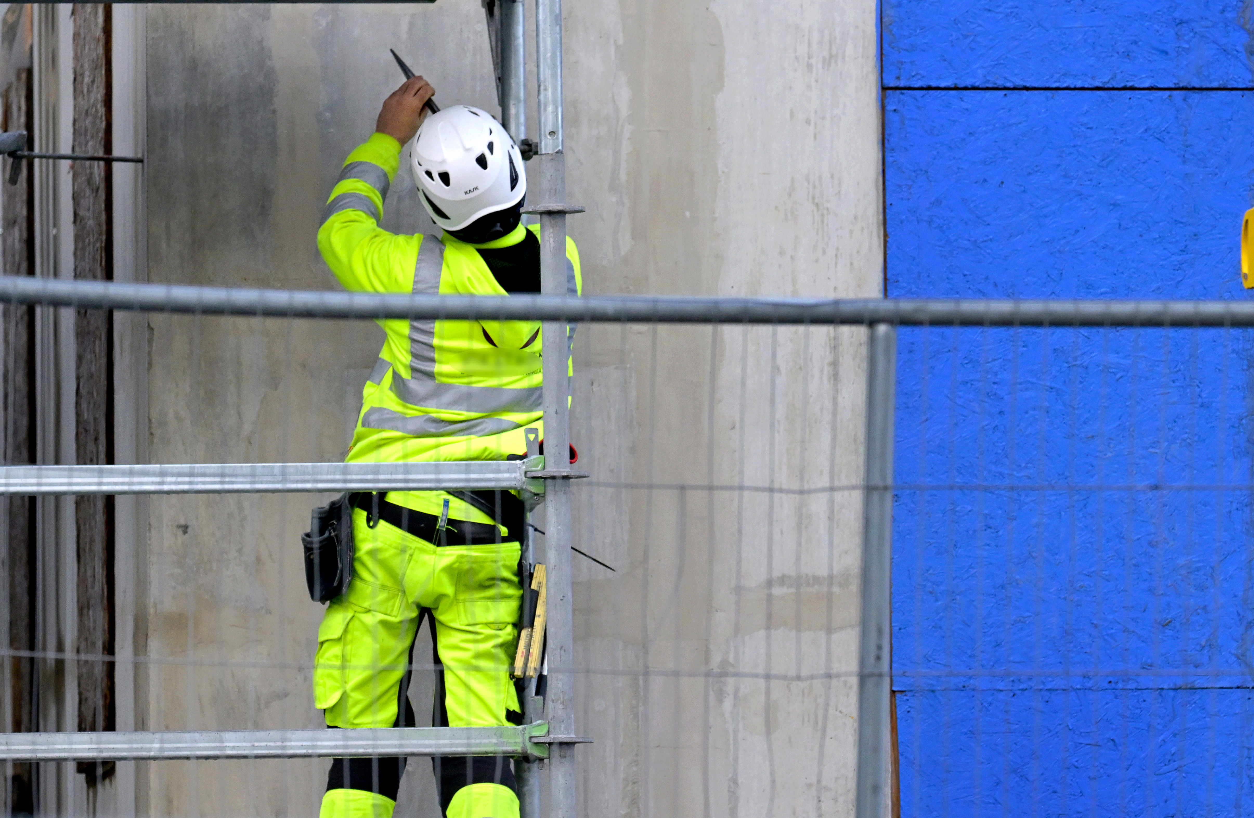 En byggarbetare iklädd skyddsutrustning och hjälm står på en byggnadsställning och målar en vägg. En blå panel syns till höger. Säkerhetsbarriärer finns i förgrunden.