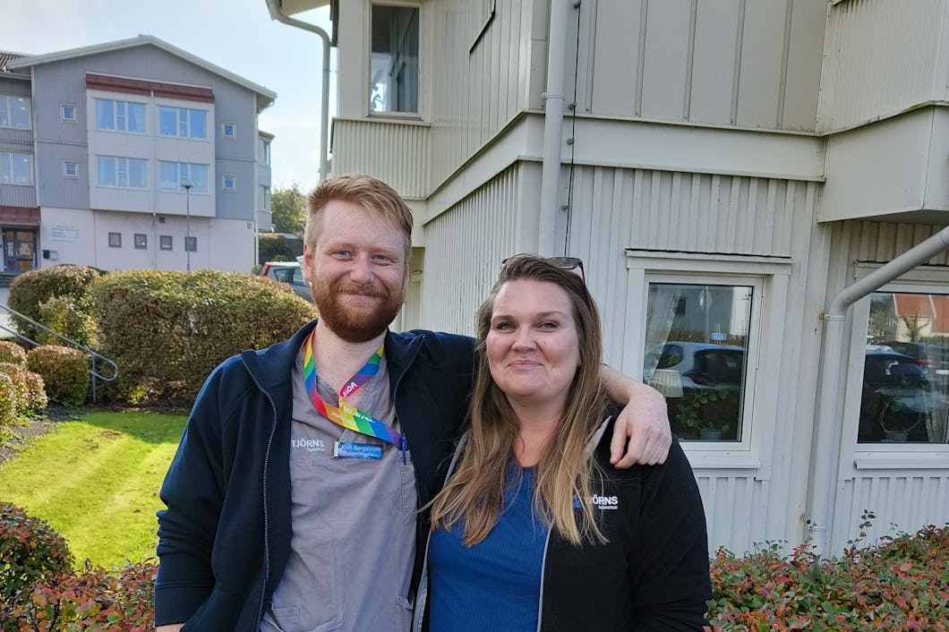Axel Bergström och Maria Zachrisson står utomhus utomhus en byggnad med vit ytterpanel. Den ena personen har en arm runt den andra. I bakgrunden syns buskar och gräs.