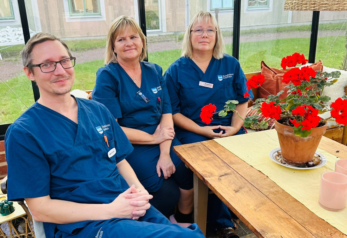 Tre personer i blå sjukhuskläder, Mathias Karlsson, Marie Carlsson och Madeleine Isaksson, sitter vid ett träbord med krukväxter med röda blommor, i ett rum med stora fönster.