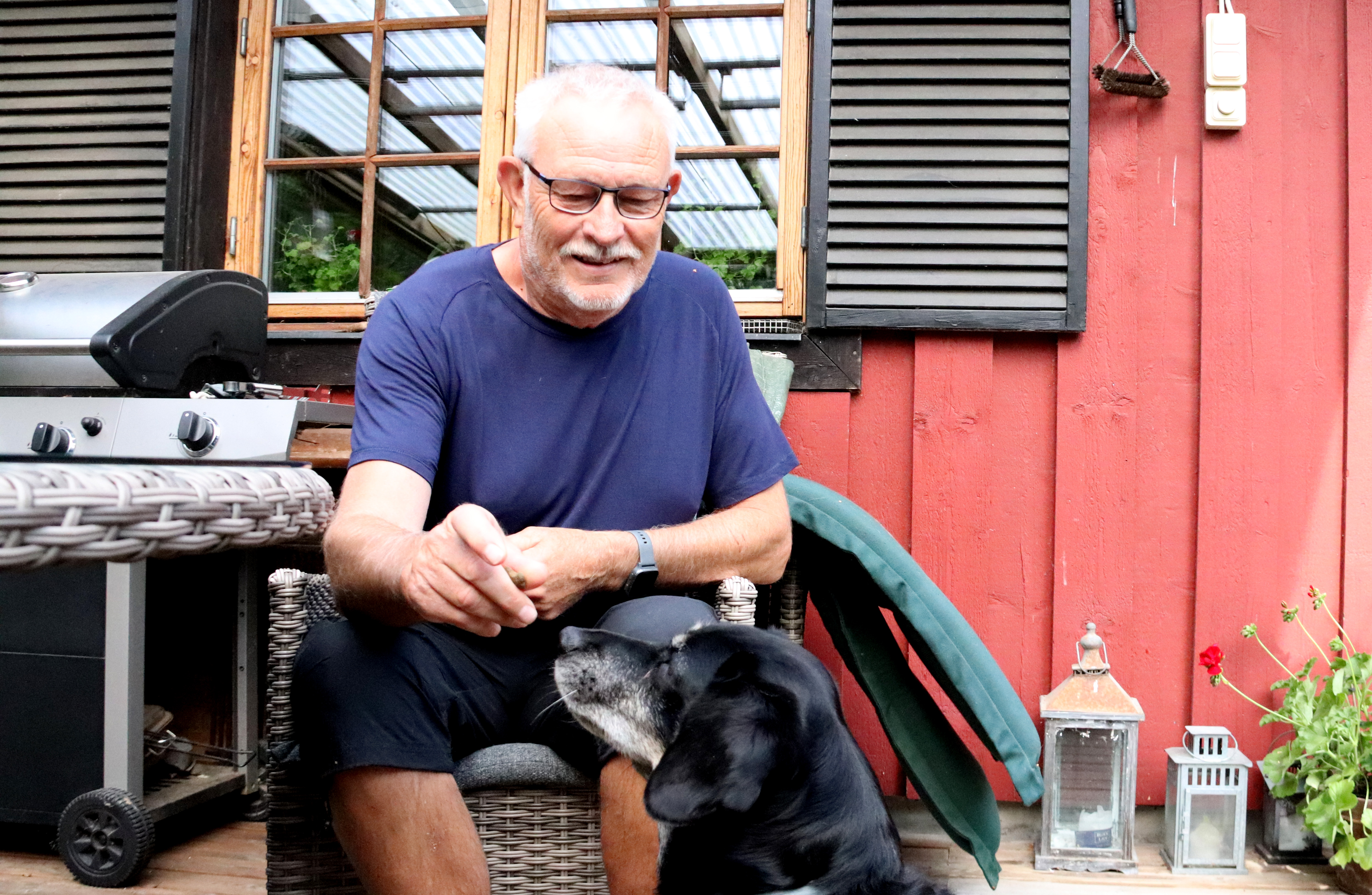 En äldre man i blå skjorta sitter på en korgstol utanför ett rött hus och ger en godbit till en svartvit hund. En grill står bredvid.