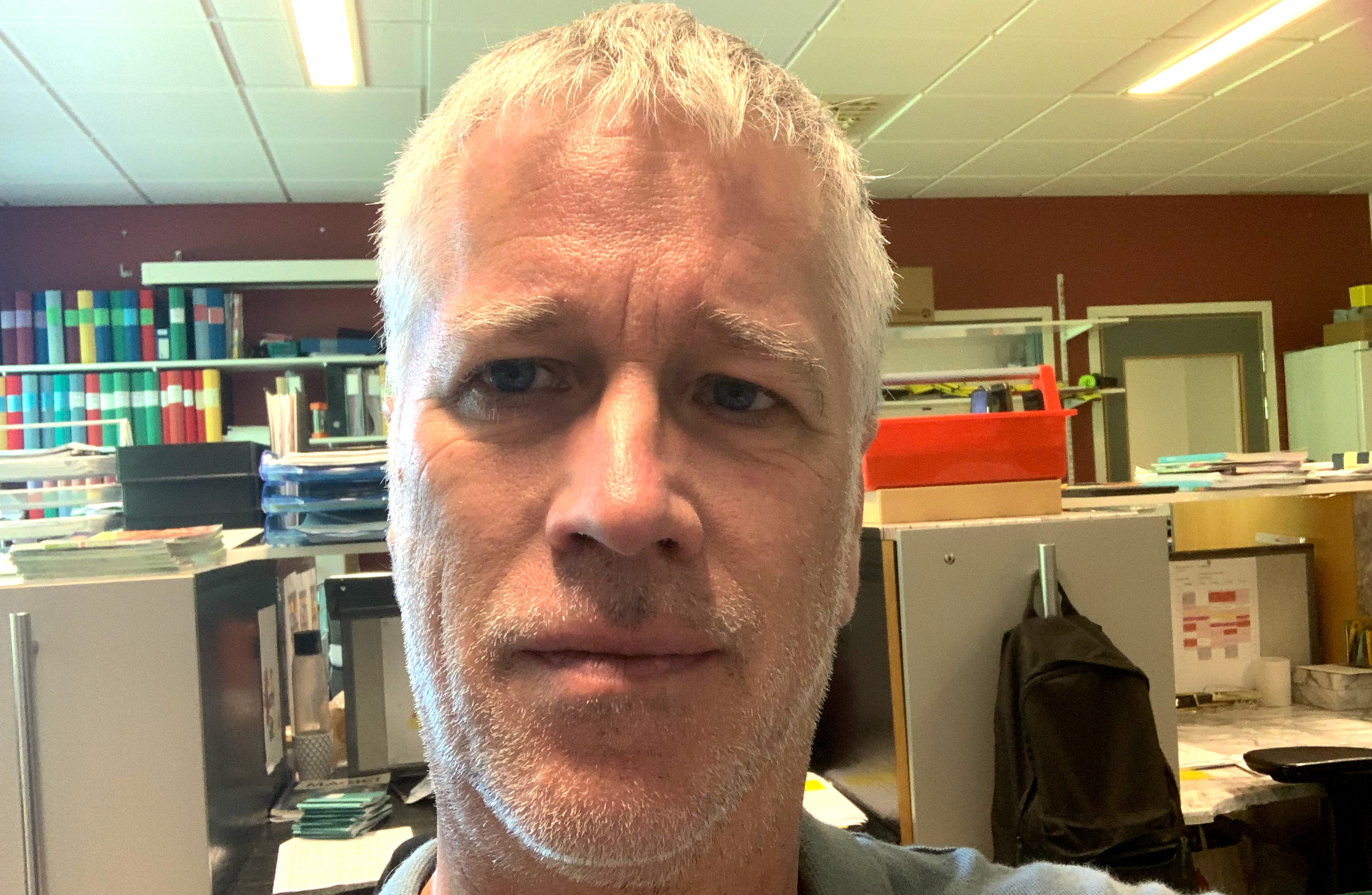 En person med kort grått hår tittar rakt mot kameran i en kontorsmiljö med hyllor, filer och lysrörsbelysning i bakgrunden.