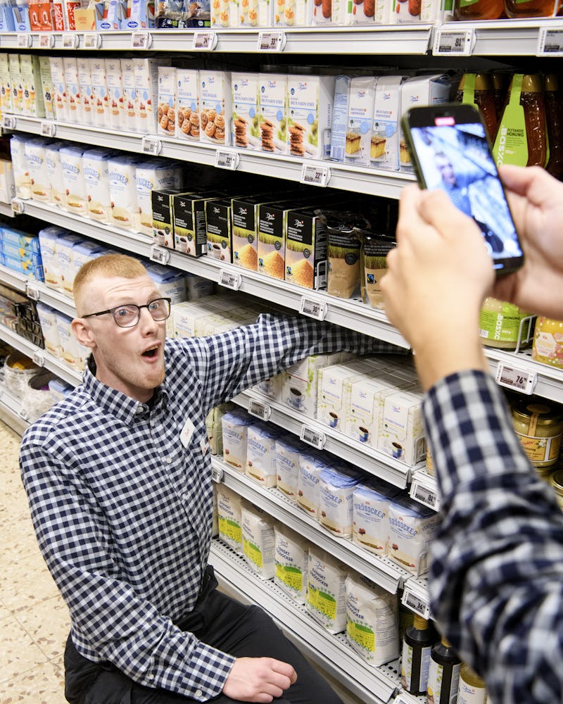 En person knästående i en gång i ett snabbköp, stirrar rakt fram. En annan person tar ett foto med en smartphone.