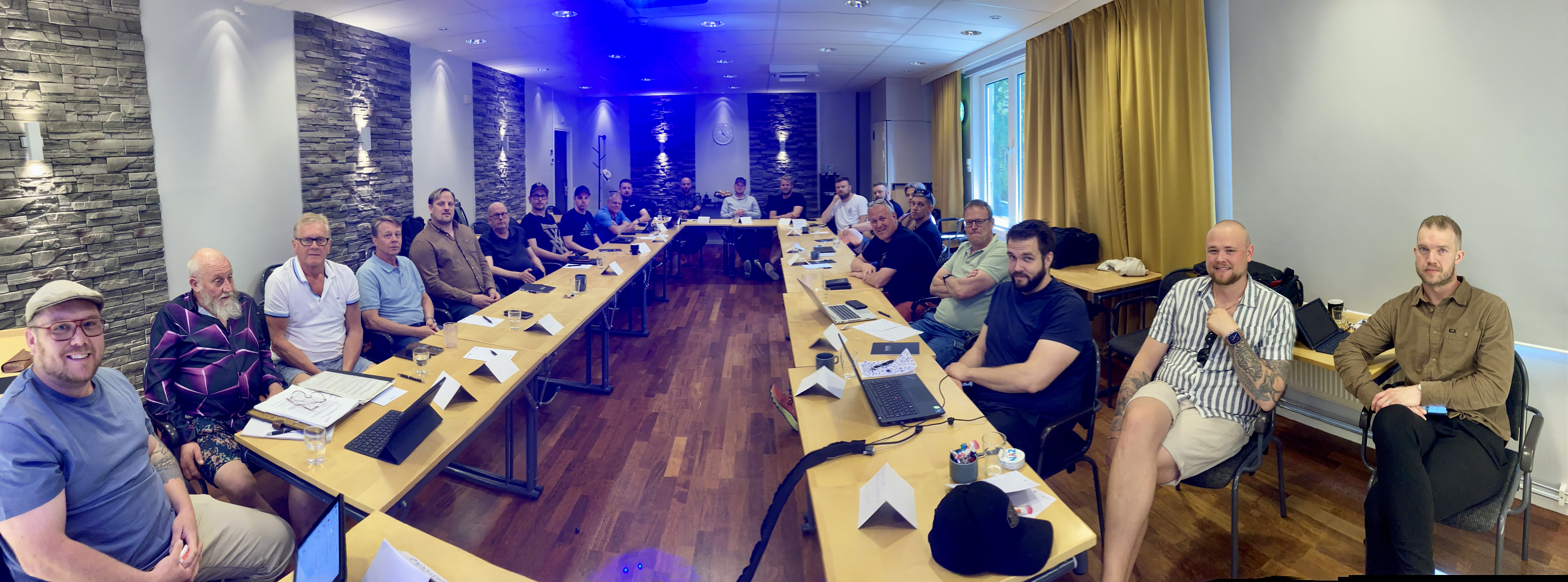 En grupp sitter människor runt ett långt rektangulärt bord i ett modernt konferensrum och deltar i ett möte.