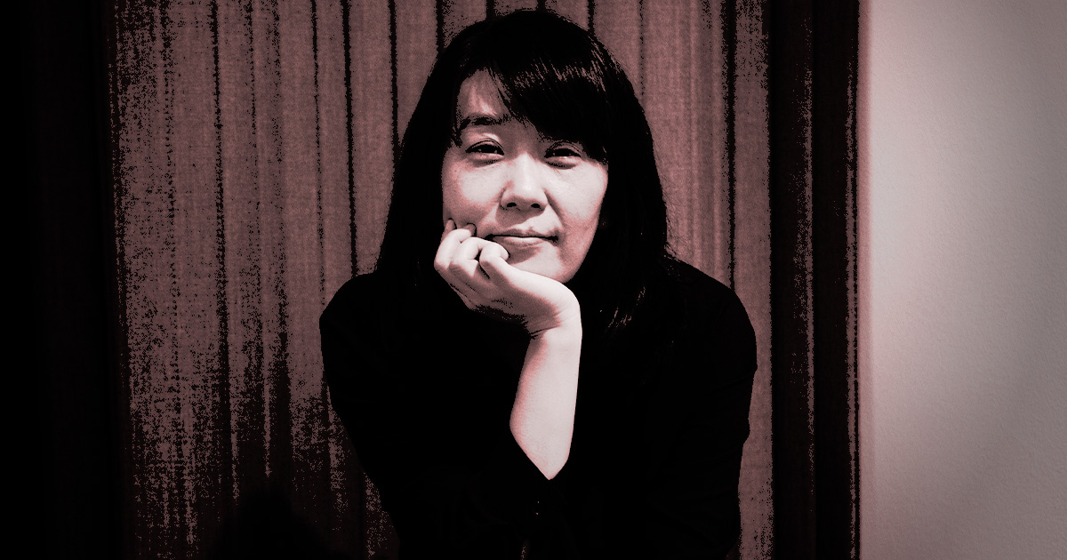 Författaren Han Kang lutar hakan mot handen och står framför stängda gardiner.