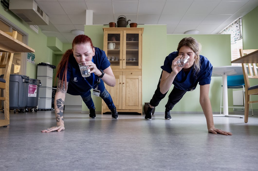 Sara Skoogh och Cecilia Käll i jobbkläder utförde plankövningar och dricker vatten i ett rum med gröna väggar och träinredning.