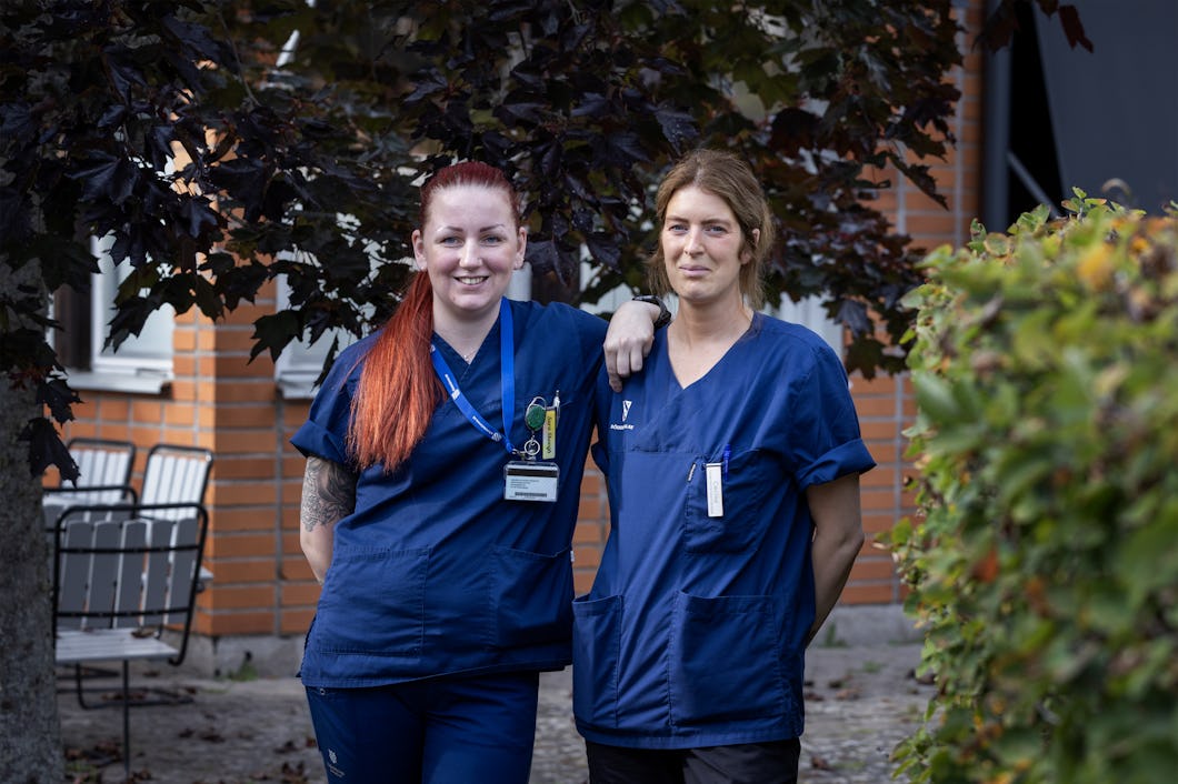 Sara Skoogh, vårdbiträde och Cecilia Käll, undersköterska vid Hemtjänsten i Söderköping i blå arbetskläder står utomhus nära en byggnad, med ens arm vilande på den andras axel.