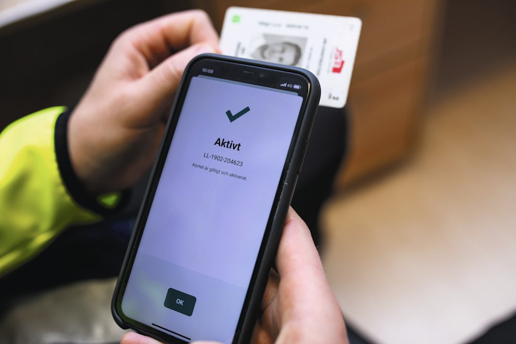 Personen håller i en smartphone som visar en skärm med ett bockmärke och texten "Aktivt", samtidigt som han håller ett ID-kort i andra handen.