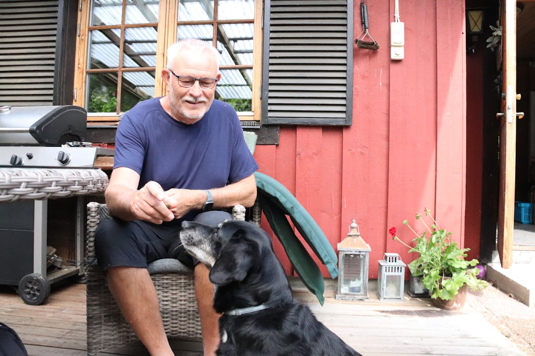 En äldre man sitter på en uteplatsstol och klappar en svart hund. I bakgrunden syns en röd stugvägg, ett bord av rotting, en grill och två lyktor.