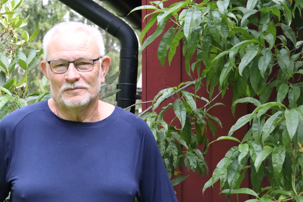 En äldre man med glasögon och en mörkblå skjorta står utomhus intill gröna växter, med en röd byggnad i bakgrunden.