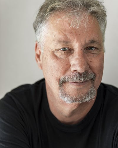 Ulf Eriksson med kort grått hår och en getskägg, klädd i en svart skjorta, tittar in i kameran med ett neutralt ansiktsuttryck.