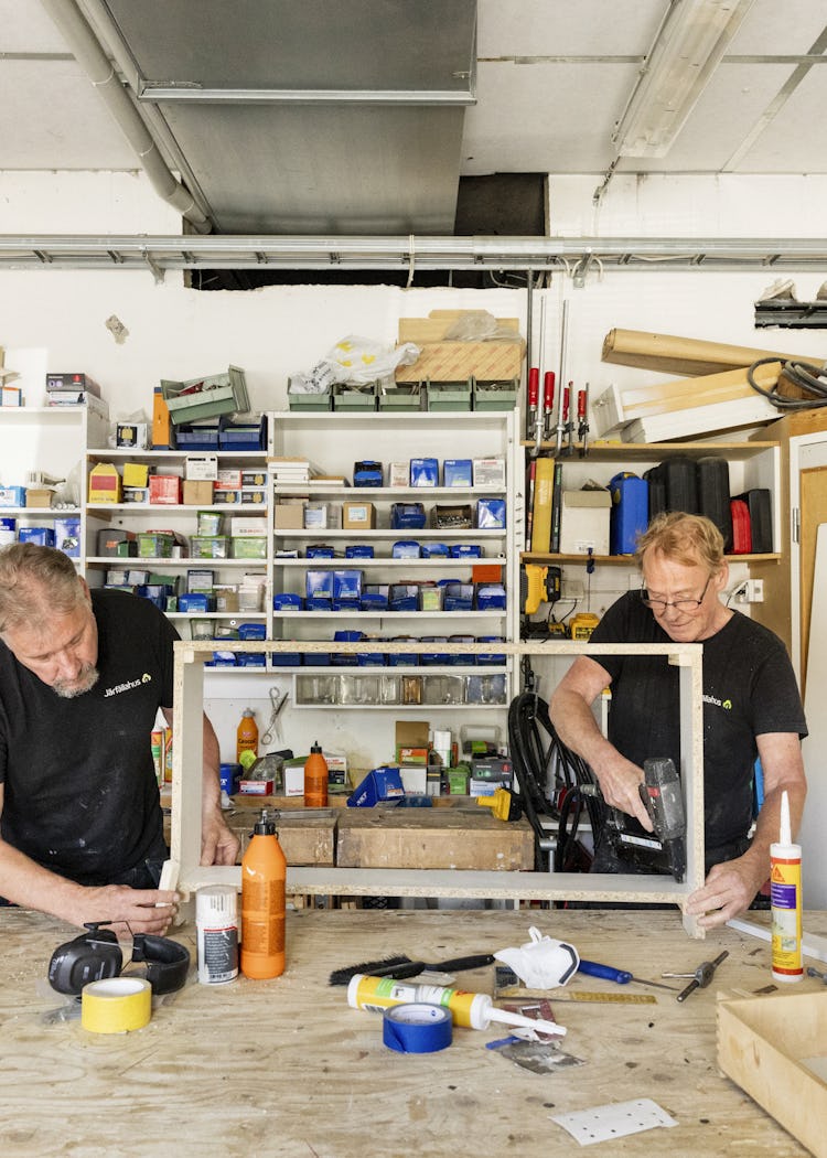 Ulf Eriksson och Göran Norr jobbar vid ett belamrat arbetsbord och monterar träramar. I bakgrunden syns hyllor fyllda med verktyg och material.