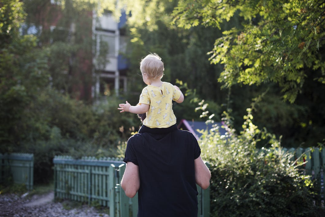 En person bär ett barn på sina axlar genom en park med mycket grönska, med ett trästaket i bakgrunden.