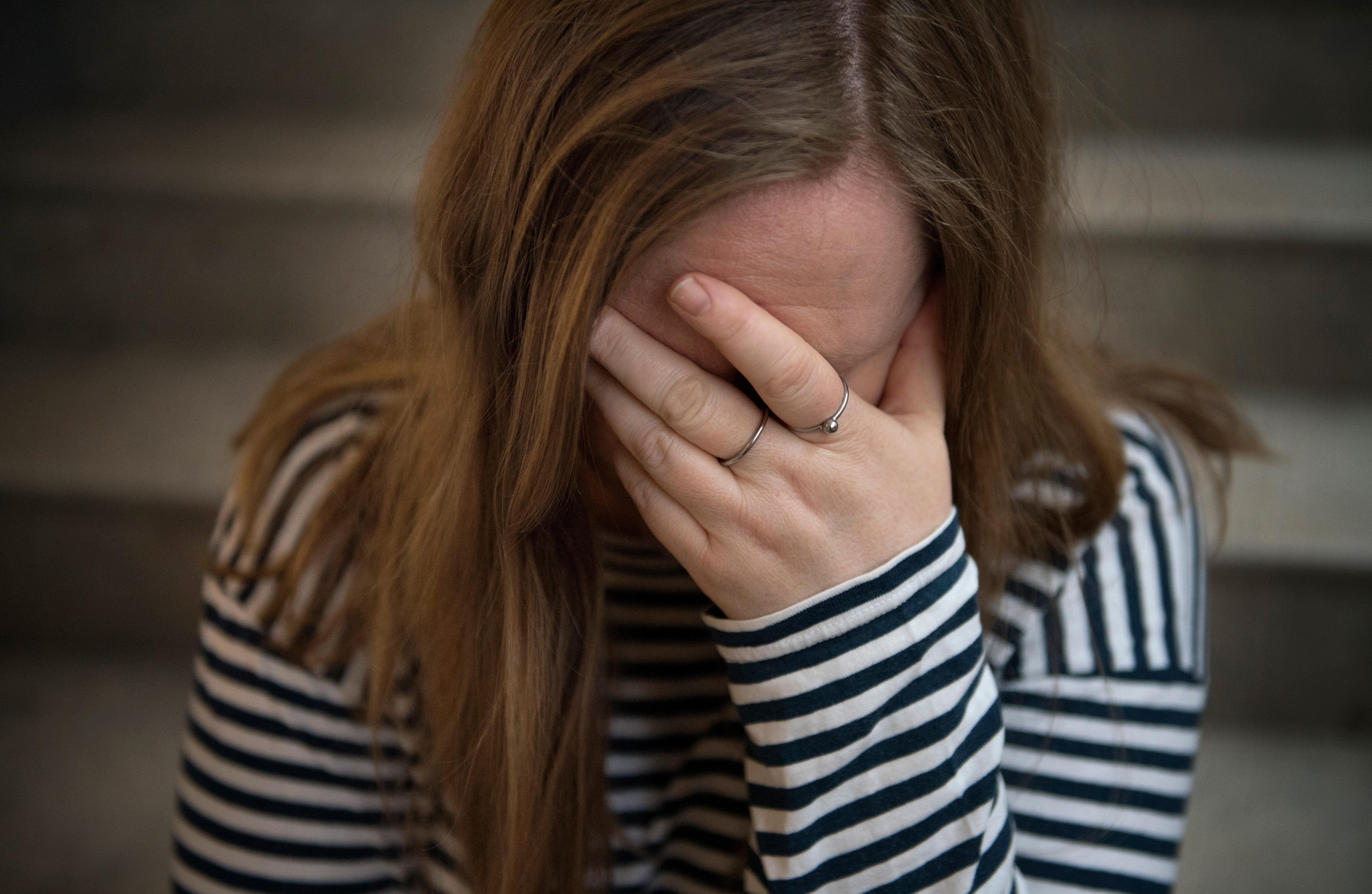 En person med långt hår i en randig tröja sitter på en trappa och täcker sitt ansikte med handen.