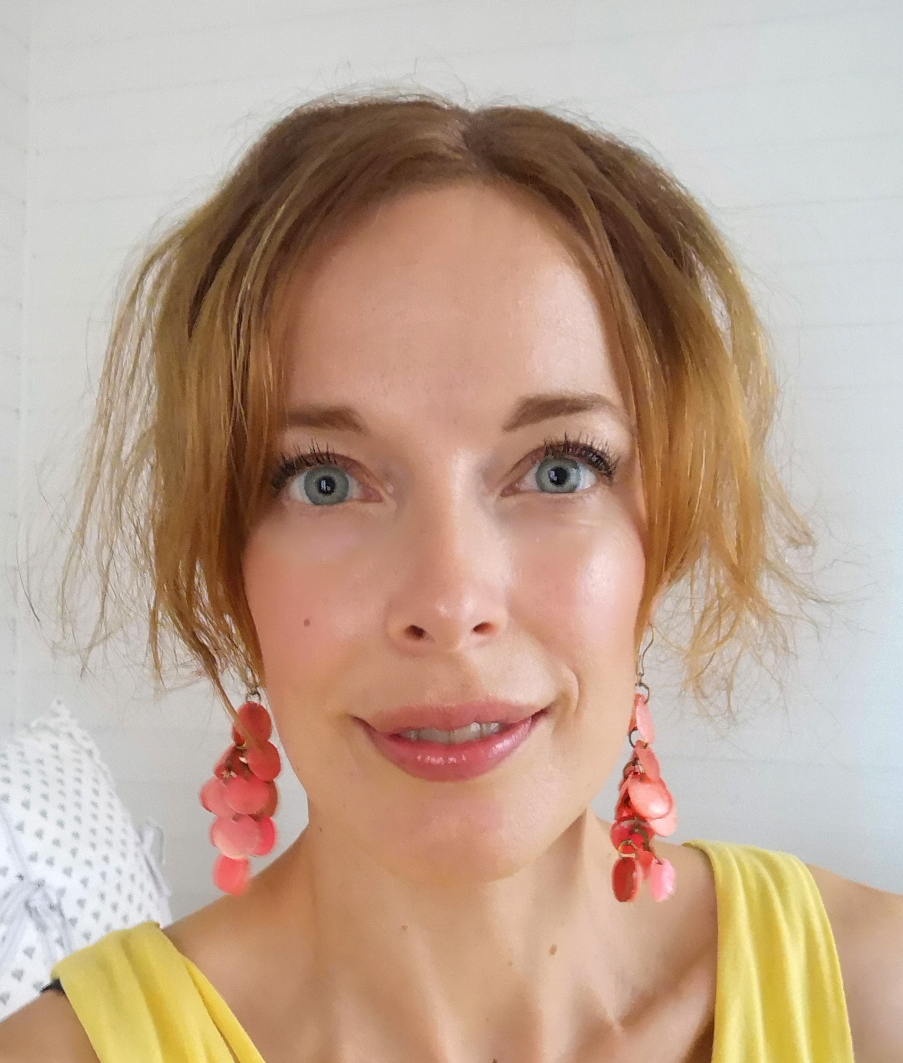 En kvinna med ljus hy och ljust brunt hår bär rosa örhängen och en gul topp. Hon ler mot kameran med en ljus bakgrund.