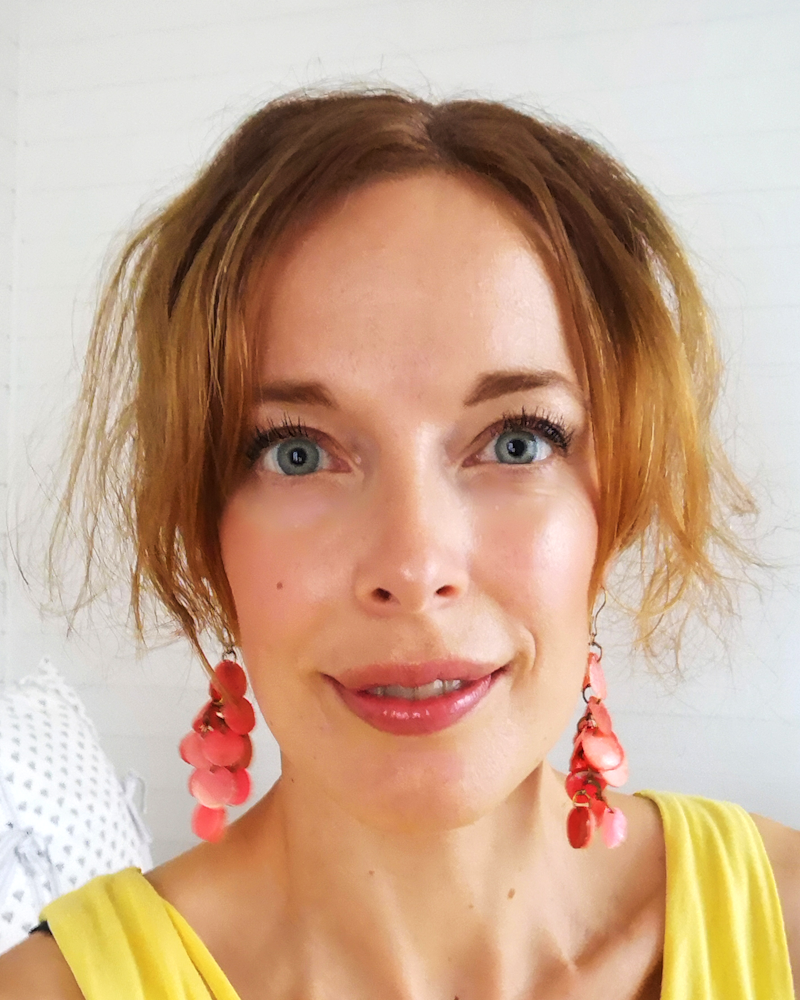 Lena Petersson med kort, vågigt brunt hår bär stora rosa örhängen och en gul topp. Personen ler mot kameran. Bakgrunden är vit och enkel.