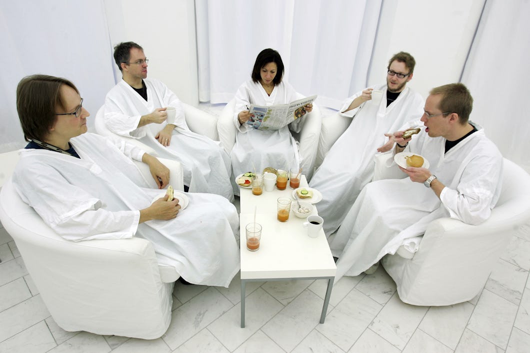 Fem personer i vita kläder sitter i ett rum med vita väggar och vit golv. De är samlade runt ett litet bord med drycker och snacks. En person läser en tidning.