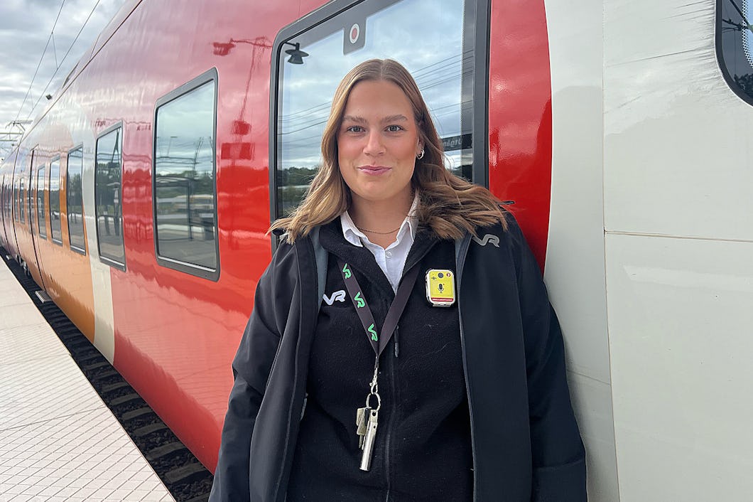 Patricia Lindström står bredvid ett rött och orange tåg från Östgötapendeln på en perrong, iförd uniform med en bricka och nycklar.