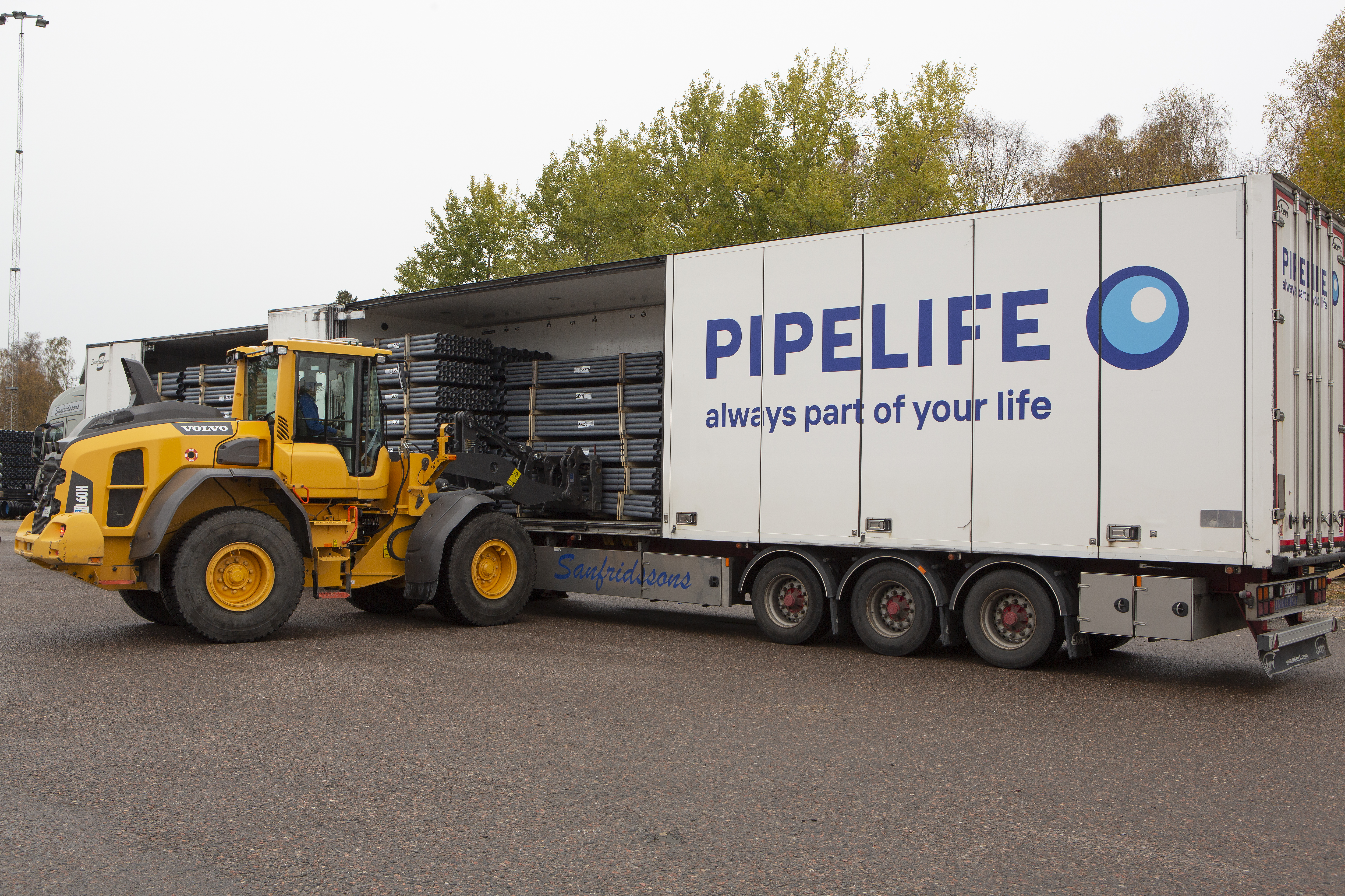 En gul lastmaskin lastar ett vitt släp med texten "PIPELIFE, always part of your life" bredvid en hög med rör.