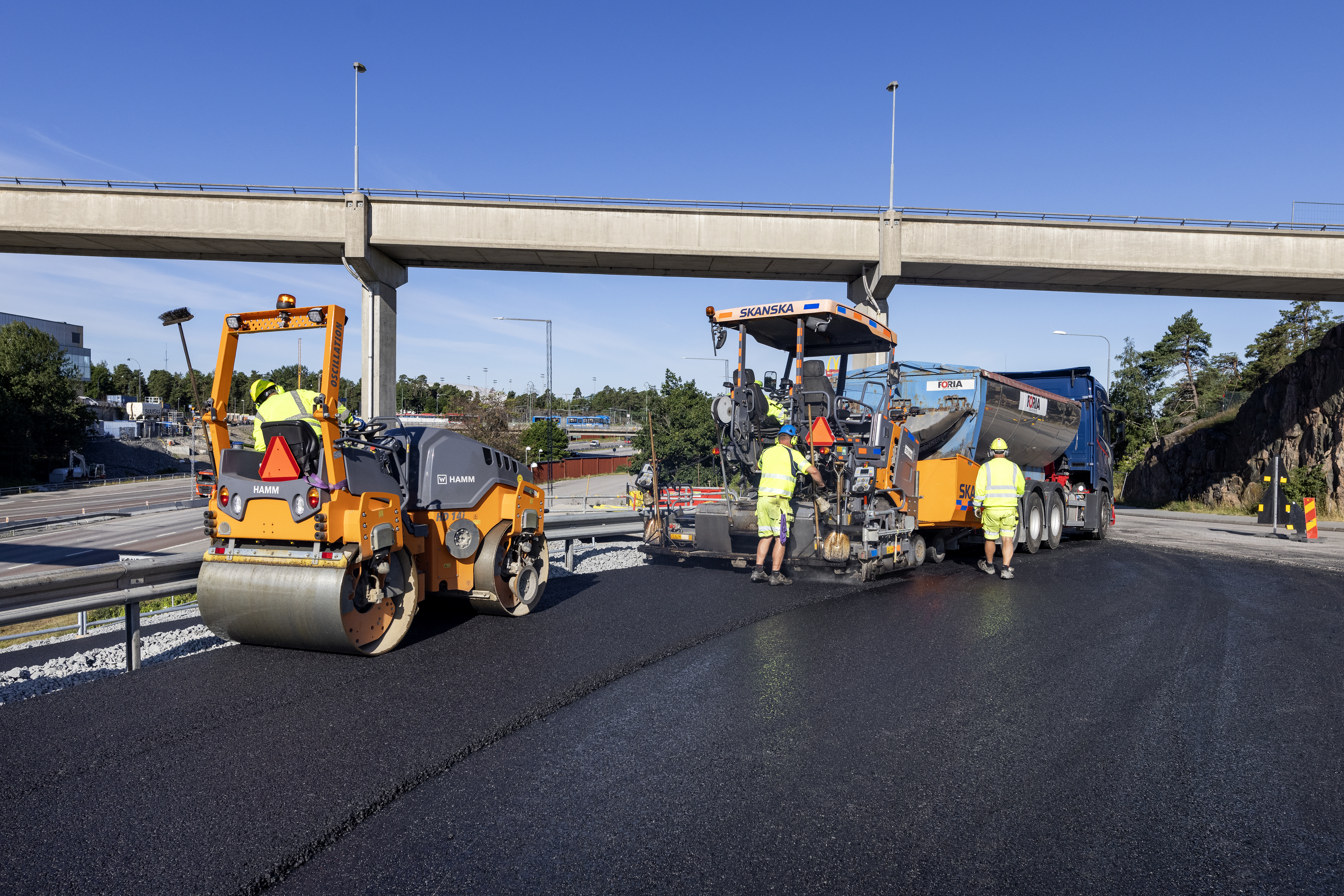 Byggarbetare asfalterar en väg med hjälp av tunga maskiner, inklusive en vält och en asfaltsläggare, under en bro.