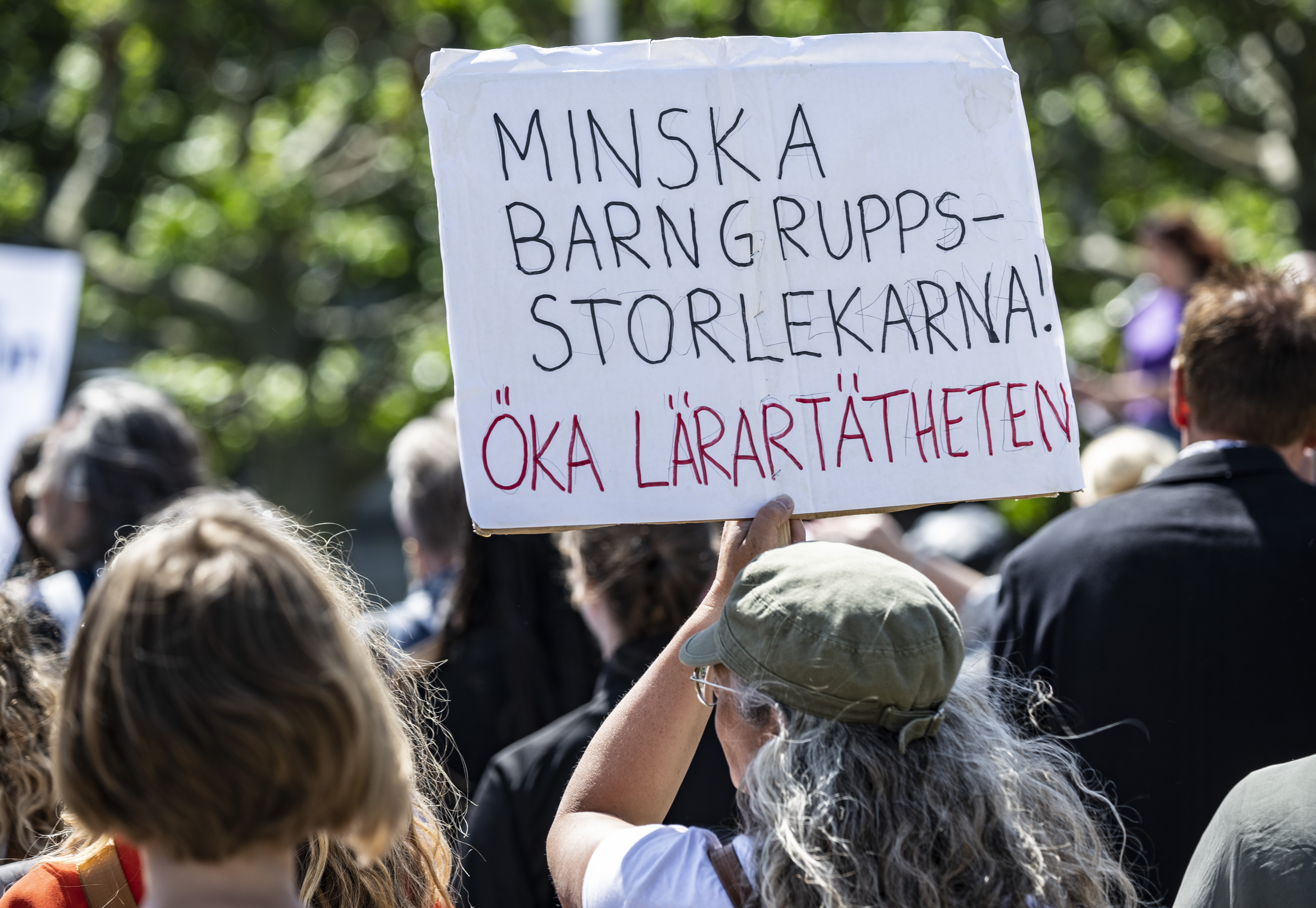 En person håller en skylt på svenska vid en offentlig sammankomst. Skylten har texten "Minska barngruppsstorlekarna! Öka lärartätheten," vilket betyder: "Minska barngruppsstorlekarna! Öka lärartätheten.