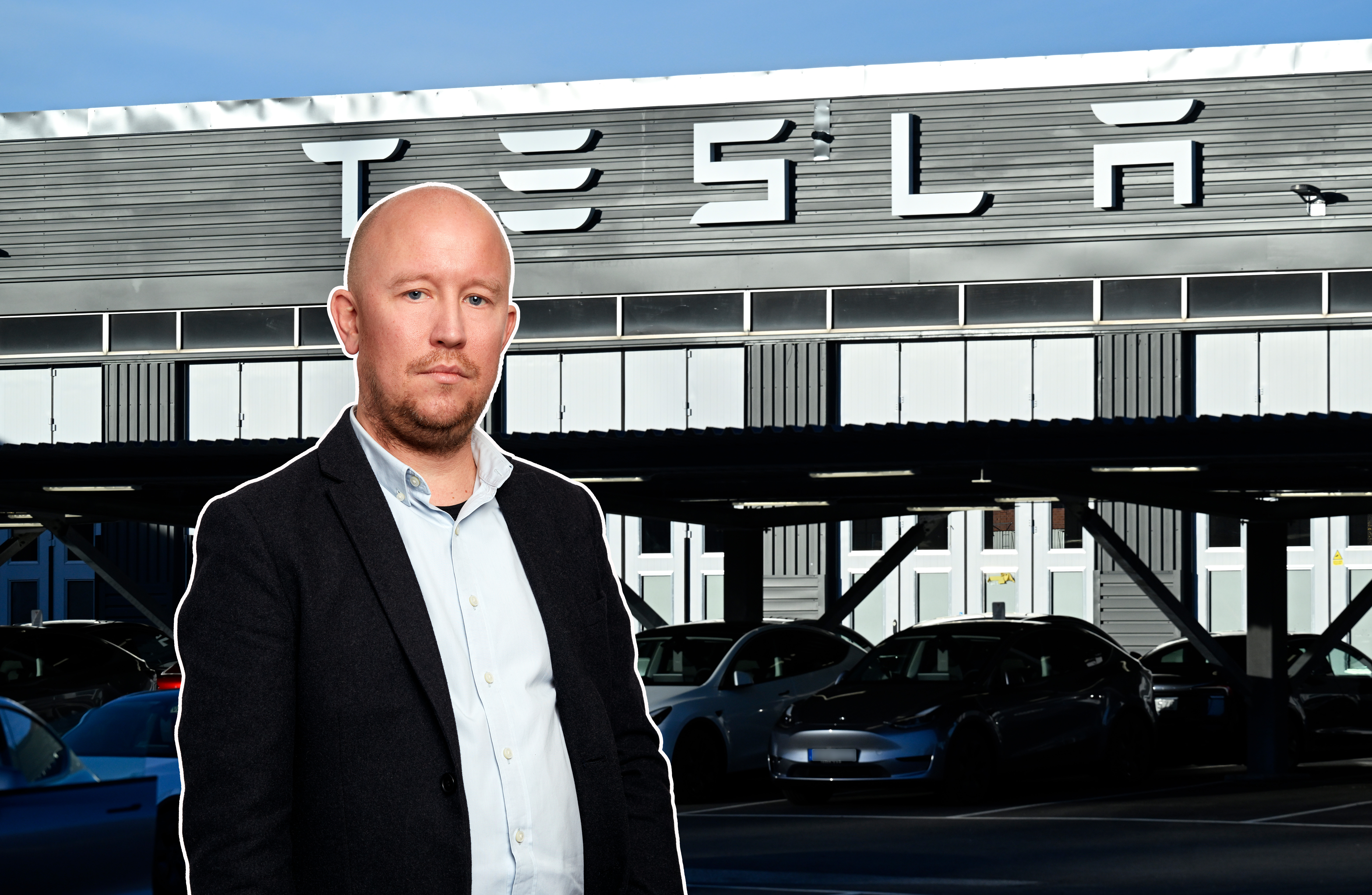 En man i kostym står framför en Tesla-byggnad med flera parkerade bilar i bakgrunden.