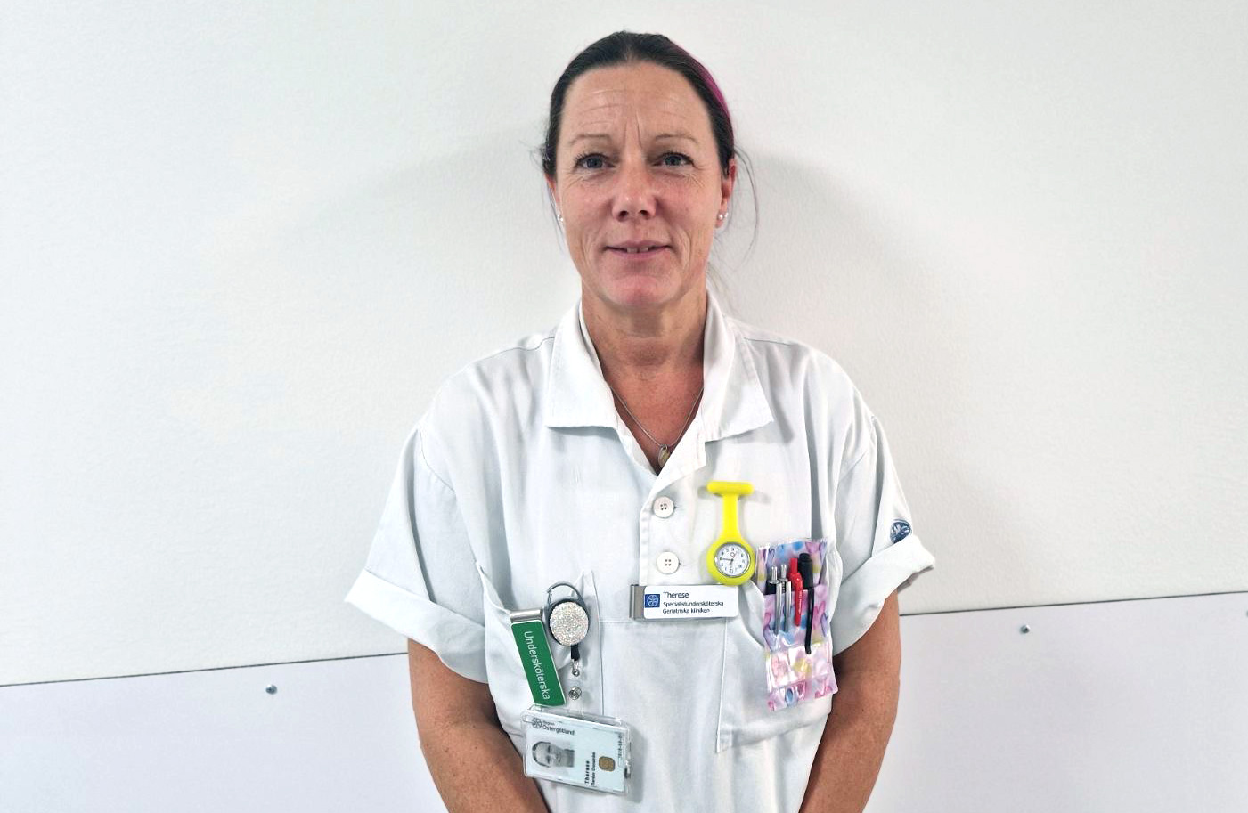 En vårdpersonal står mot en vit vägg, iklädd en vit uniform med olika verktyg och namnskyltar fästa.