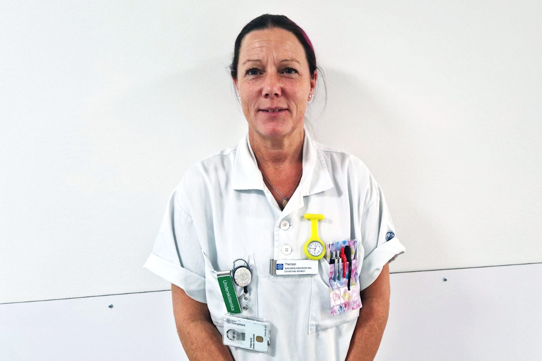 Therese Bardun Cronström. i vit medicinsk uniform står mot en ljus vägg, iklädd en namnbricka med brickor och en fastklämd klocka.