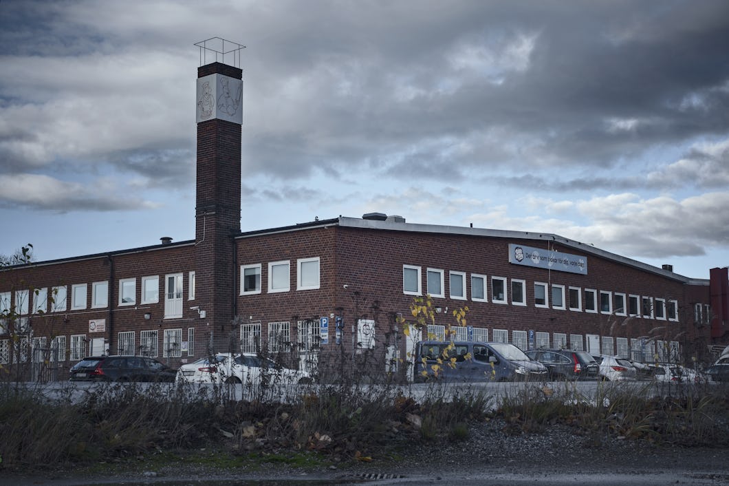 En tvåvånings industribyggnad i tegel med en högt skorsten, omgiven av parkerade bilar och molnigt väder.