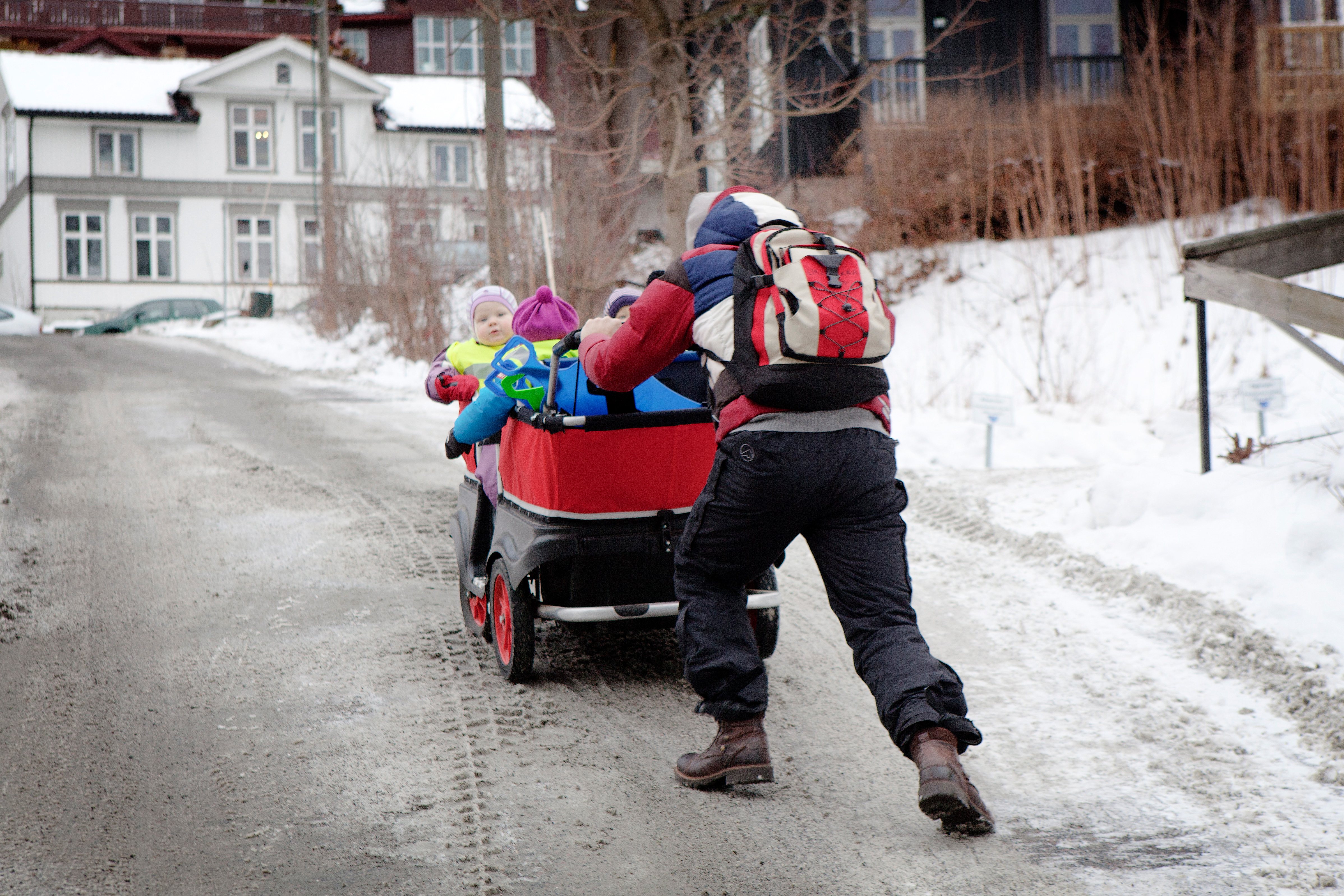 En person skjuter en röd vagn med barn i uppför en snötäckt slutande gata, med hus i bakgrunden.