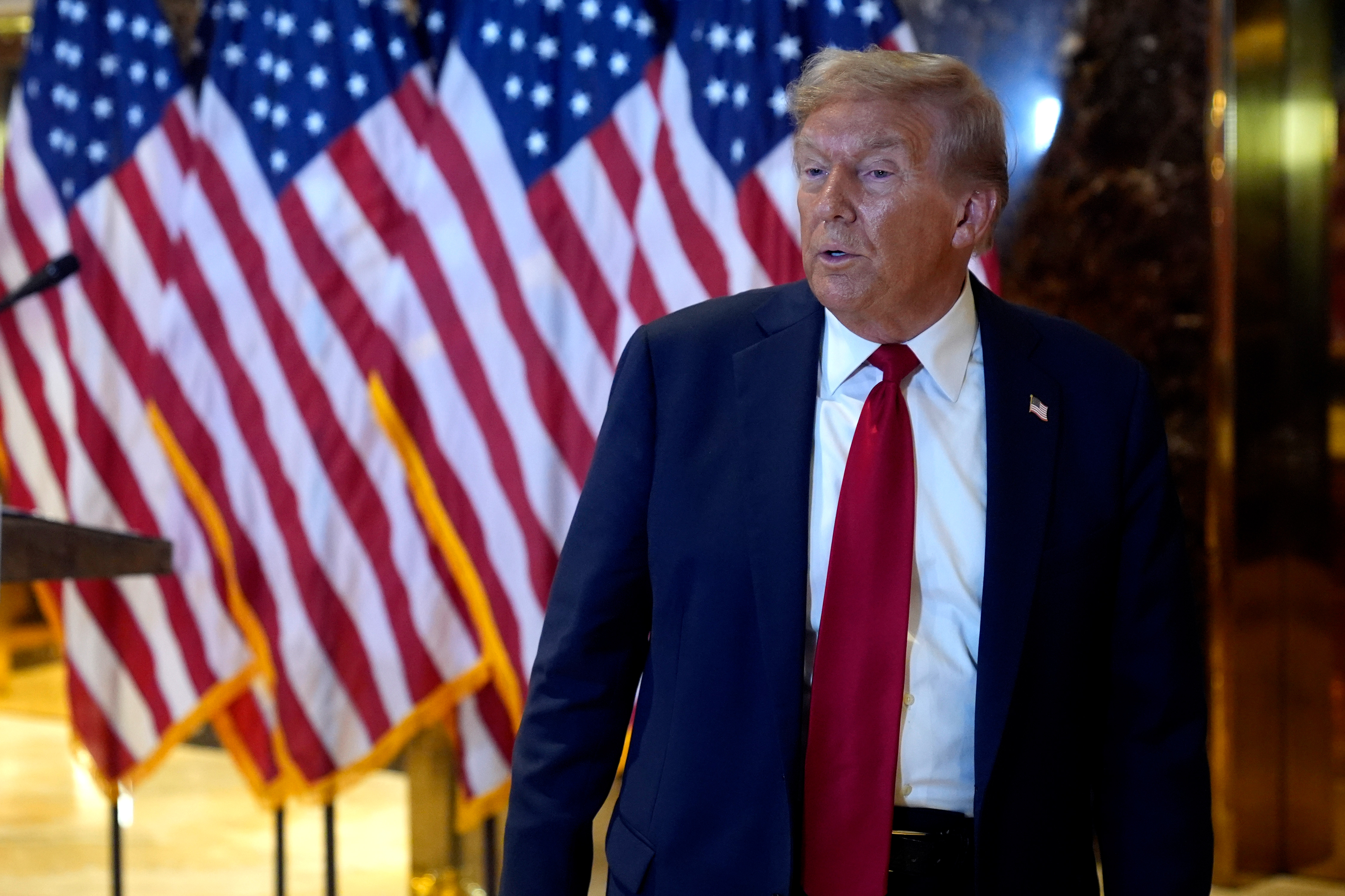 En man i kostym och röd slips inomhus, med flera amerikanska flaggor i bakgrunden.