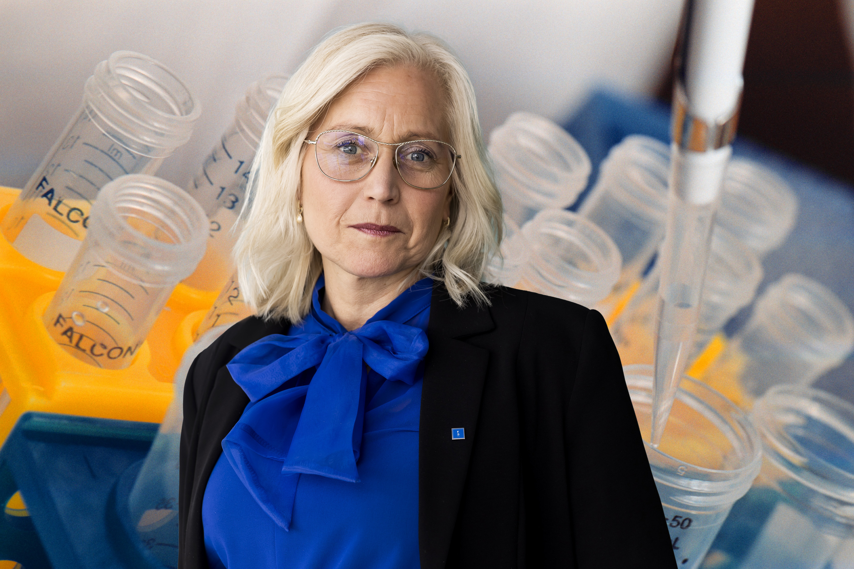 En person med ljust hår och glasögon står framför laboratorieutrustning med provrör och en pipett.