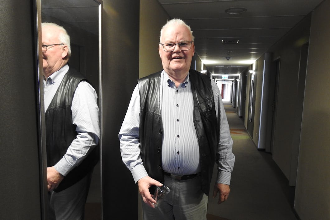 Äldre man med glasögon och läderväst står i en hall, speglas i en spegel.