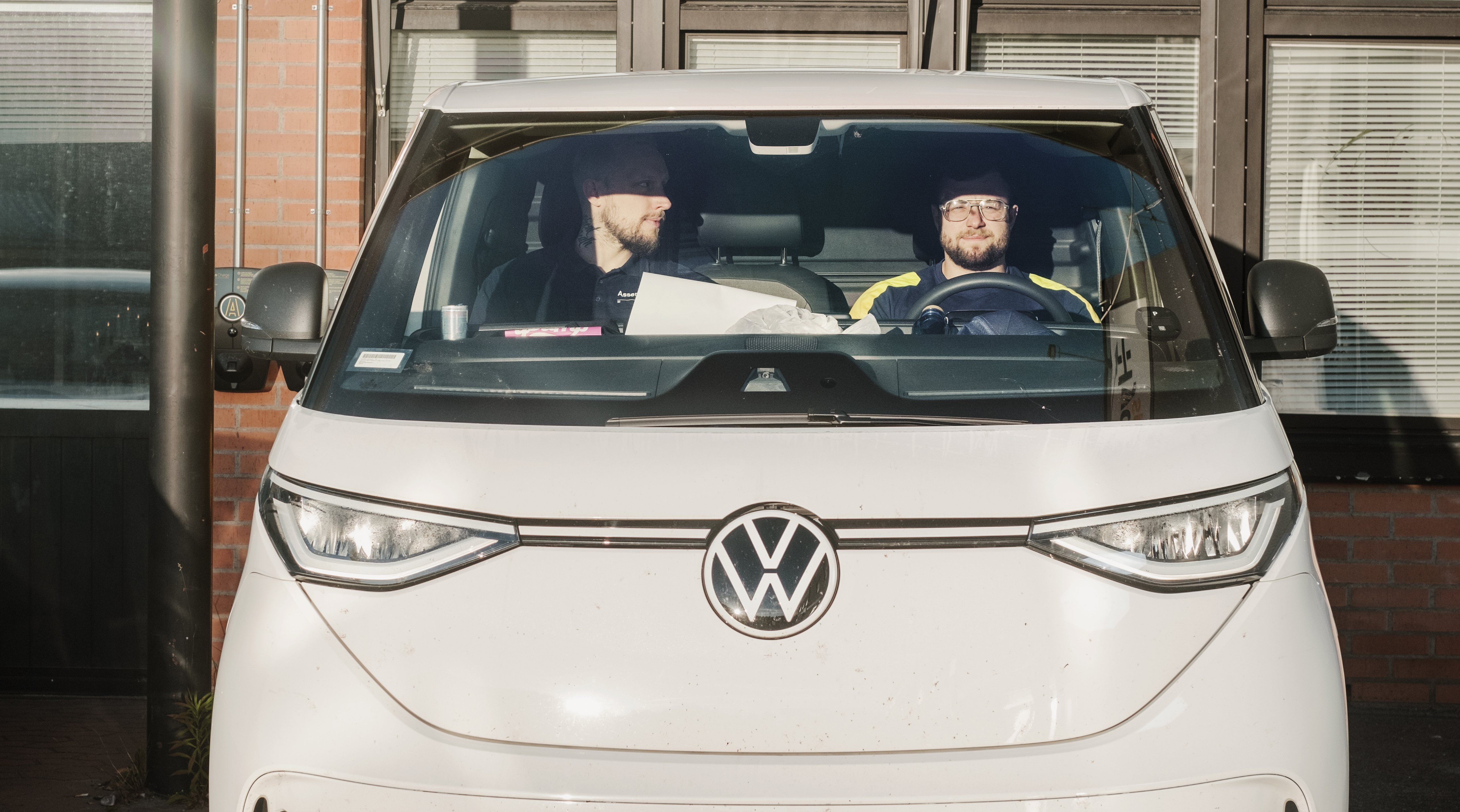 Två personer sitter i framsätena på en Volkswagen-buss som står parkerad framför en byggnad.