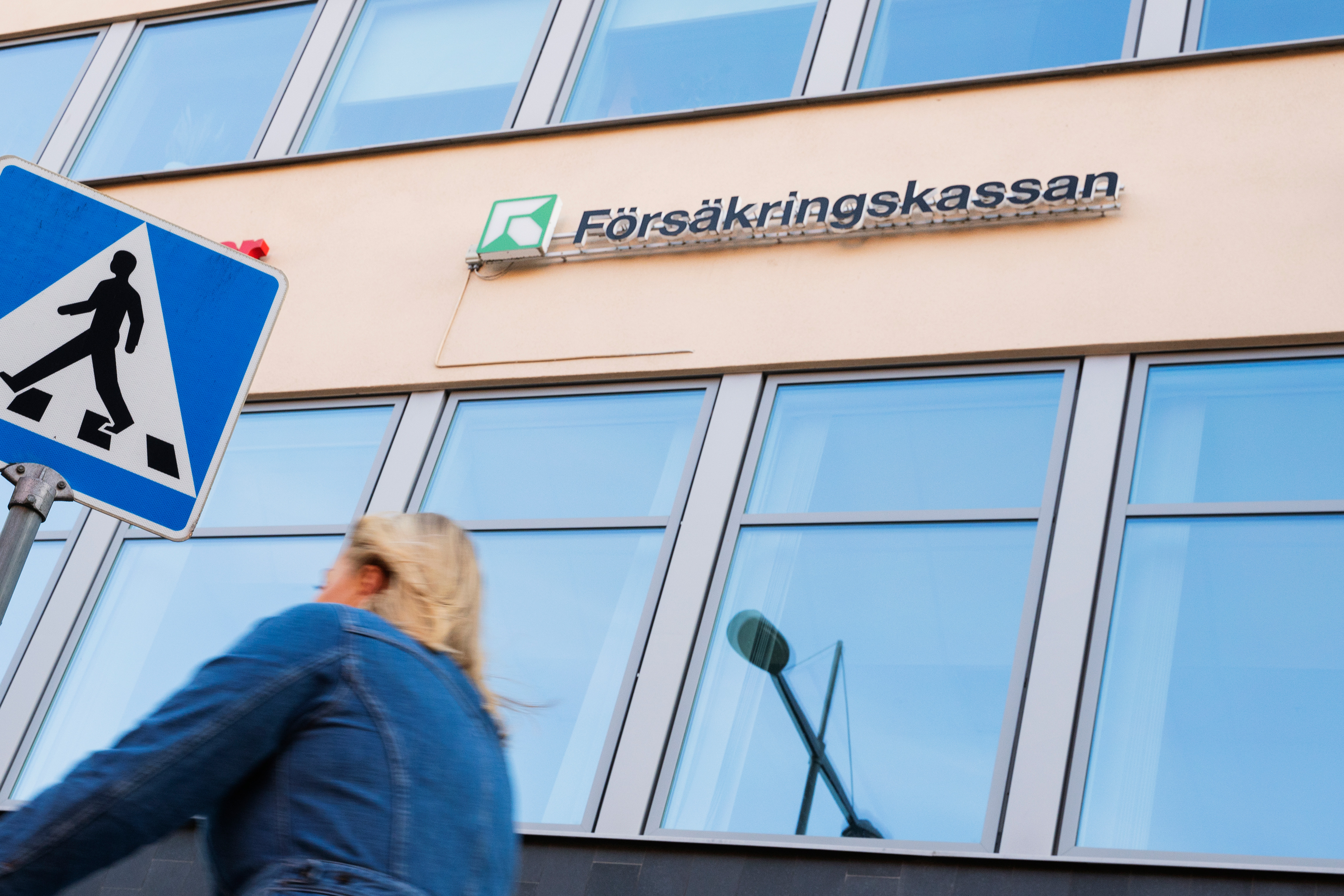 En kvinna cyklar förbi en byggnad med stora fönster. En skylt med texten "Försäkringskassan" syns ovanför. Ett övergångsställe är markerat i förgrunden.
