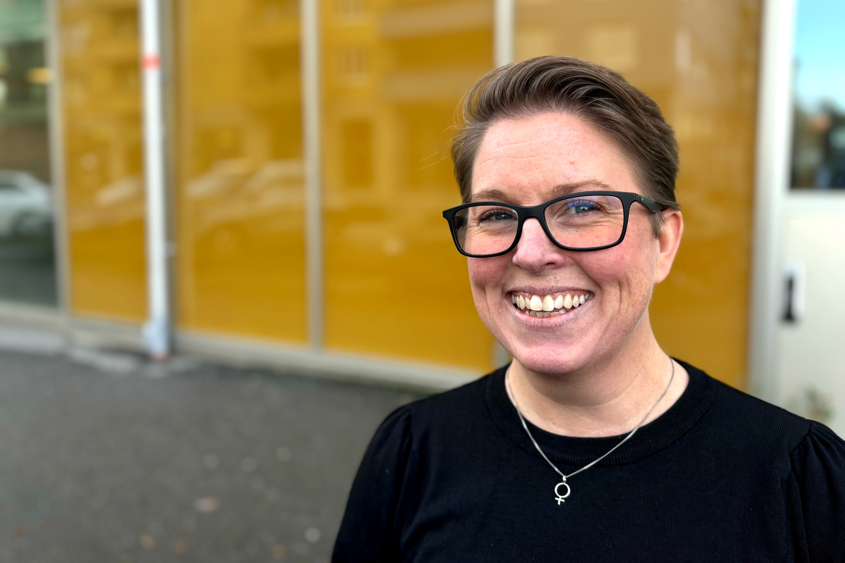 Person med kort hår och glasögon står framför en gul vägg.