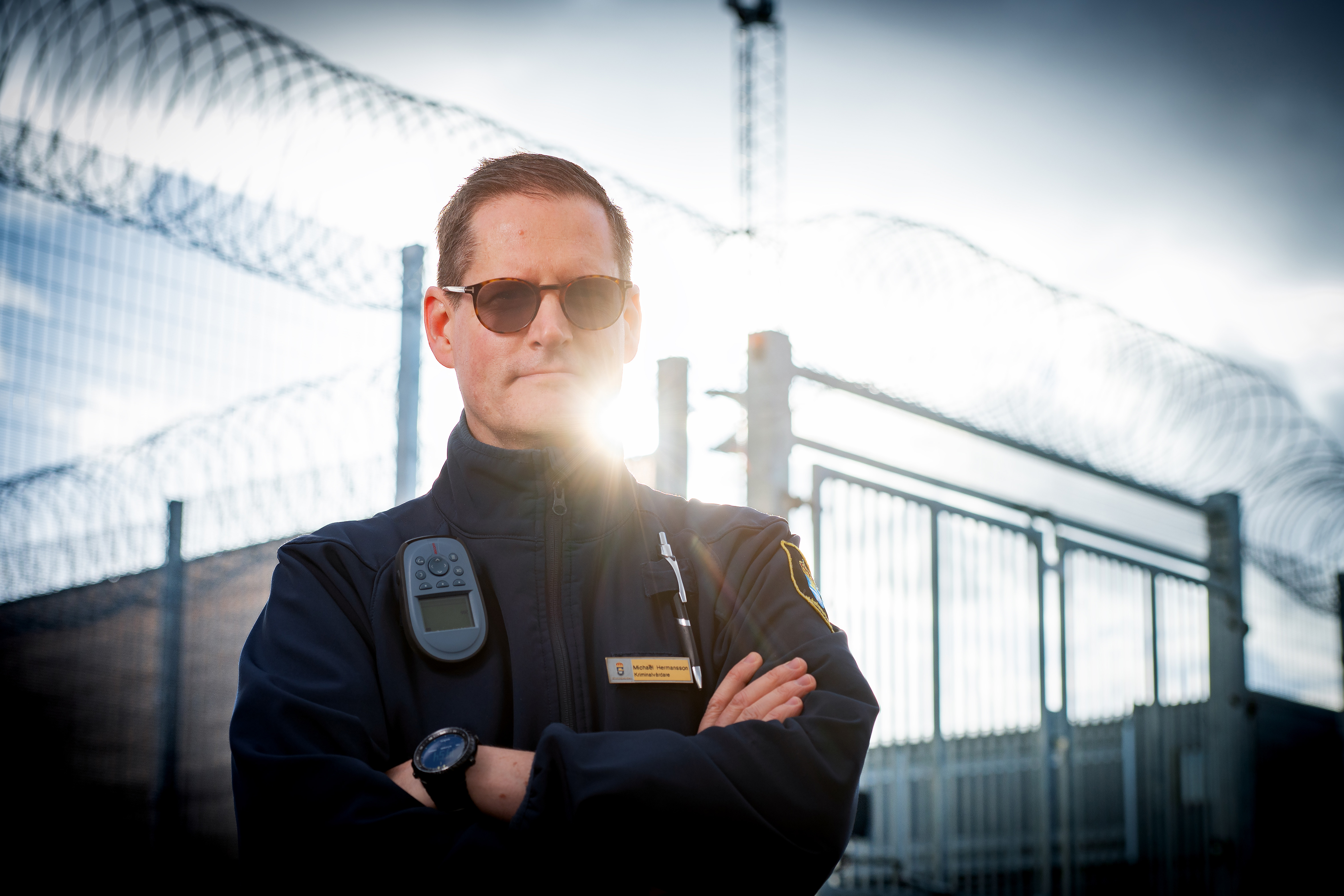 En person bär solglasögon och en mörk jacka medan de står utomhus med solljus i bakgrunden.