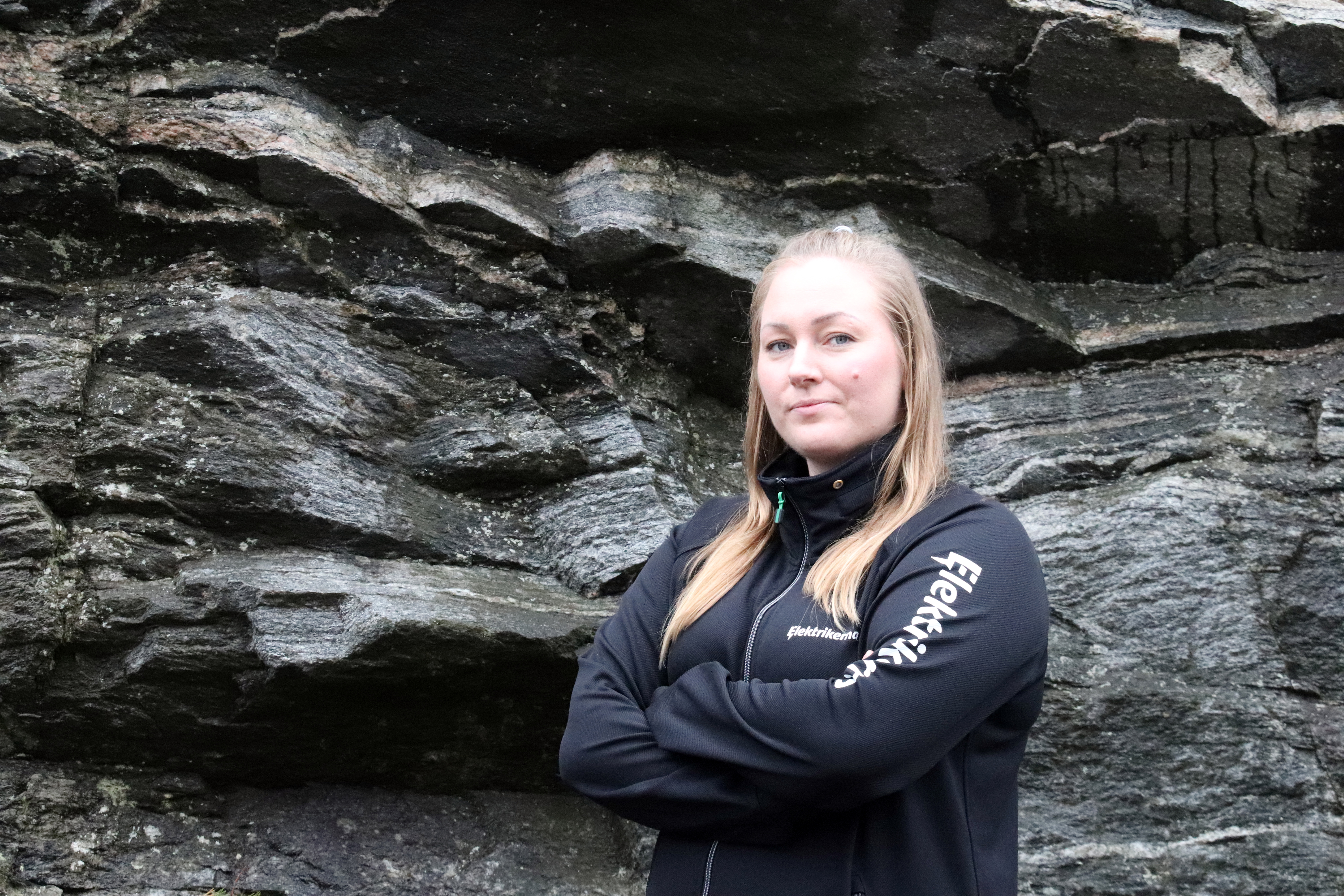 En person i svart jacka står med armarna i kors framför en vägg av skiktade stenar.