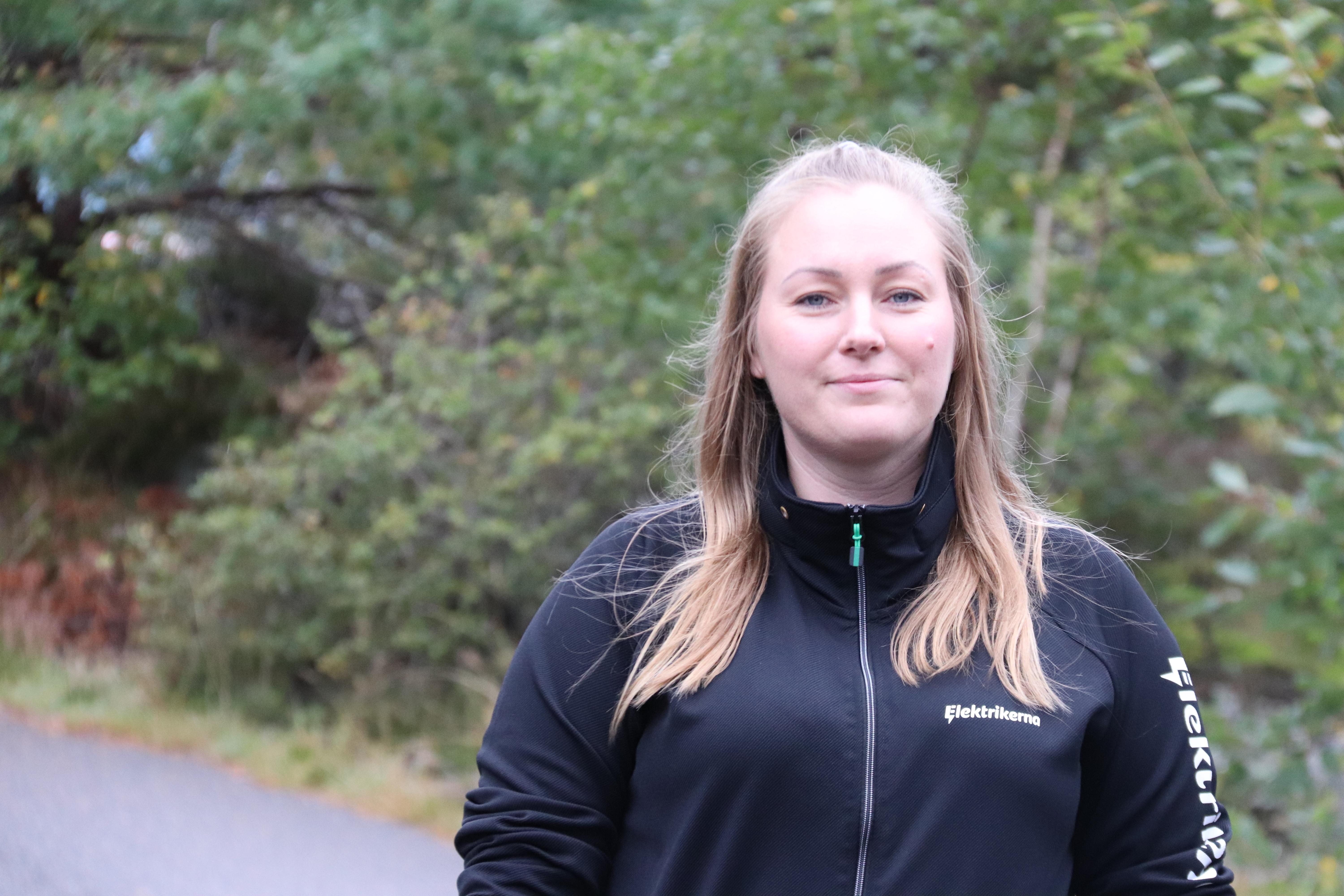 En person i svart jacka står utomhus med grönska i bakgrunden.