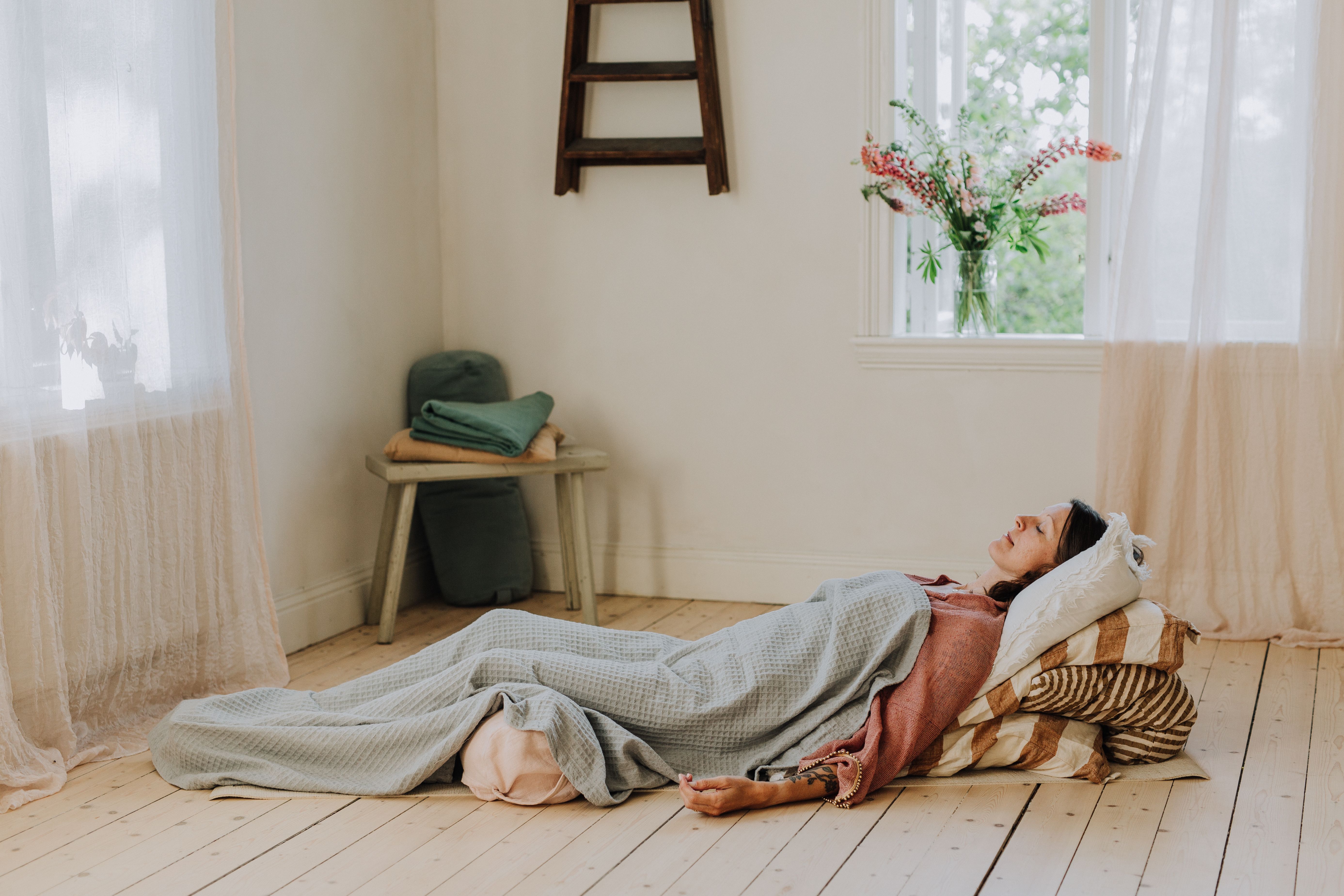 En kvinna ligger på en vadderad matta, insvept i en filt, med slutna ögon i ett solbelyst rum.