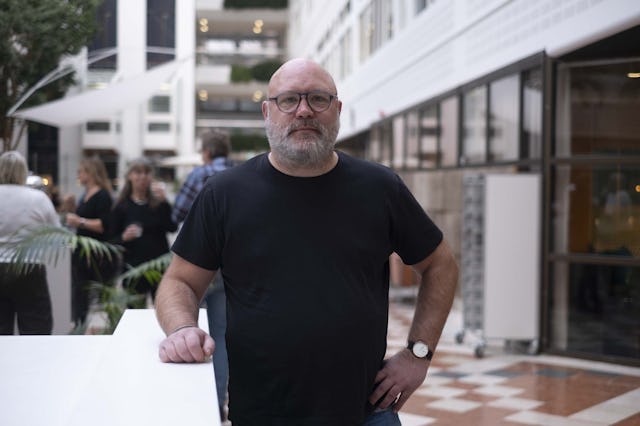 Johan Claesson med skägg och svart t-shirt står inomhus, med personer i bakgrunden.