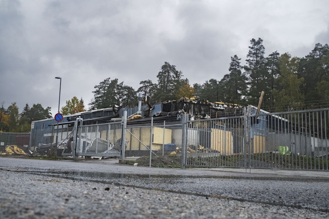 En bränd byggnad med förkolnade rester, omgiven av ett kedjelänkstängsel, med träd i bakgrunden.