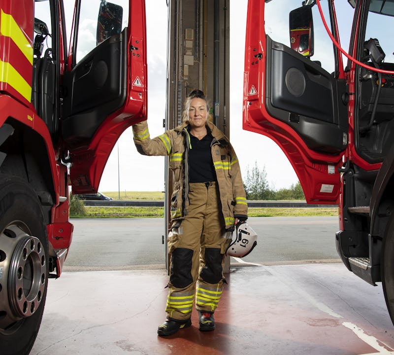 Jessica Lidberg i full utrustning står mellan två brandbilar med öppna dörrar och håller en hjälm i garaget.