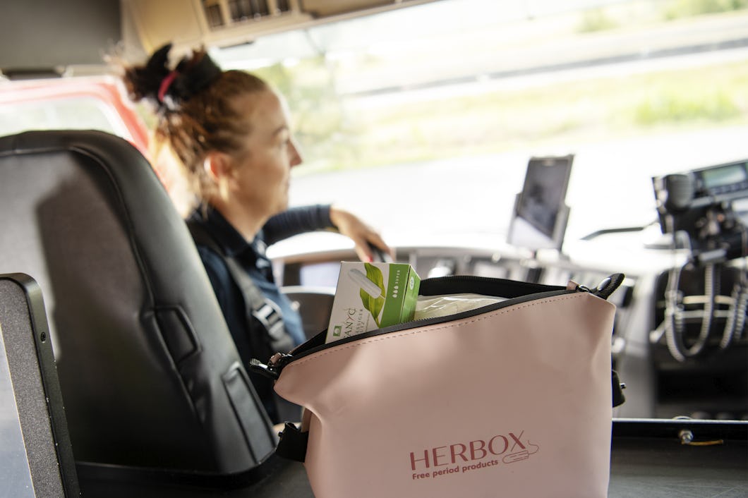 En rosa påse med etiketten "HERBOX" ligger på instrumentbrädan i en buss och innehåller gratis mensskydd. Jessica Lidberg syns i förarsätet.