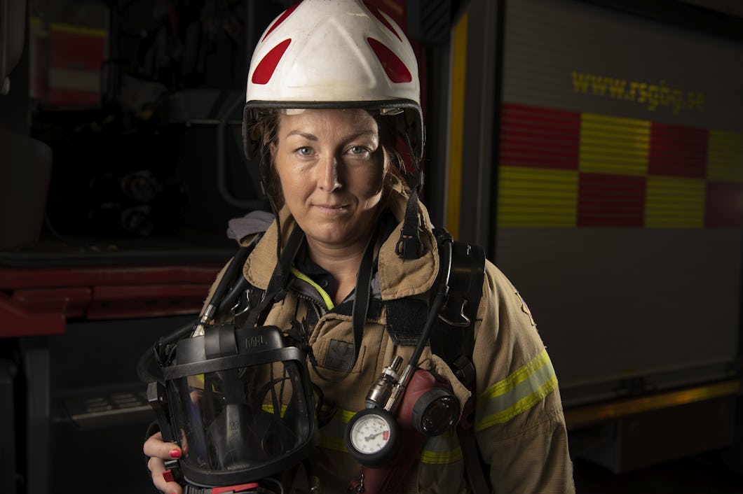 Brandmannen Jessica Lidberg i utrustning håller en hjälm och ett andningsskydd framför en brandbil.