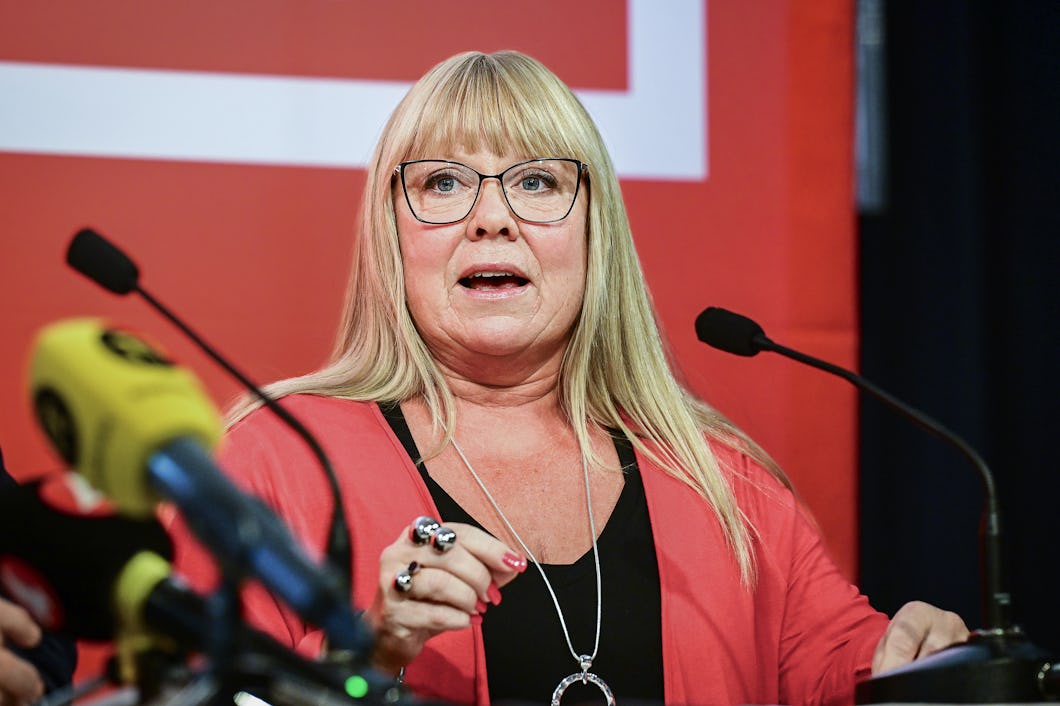 En kvinna med glasögon talar vid ett podium med mikrofoner framför en röd bakgrund.