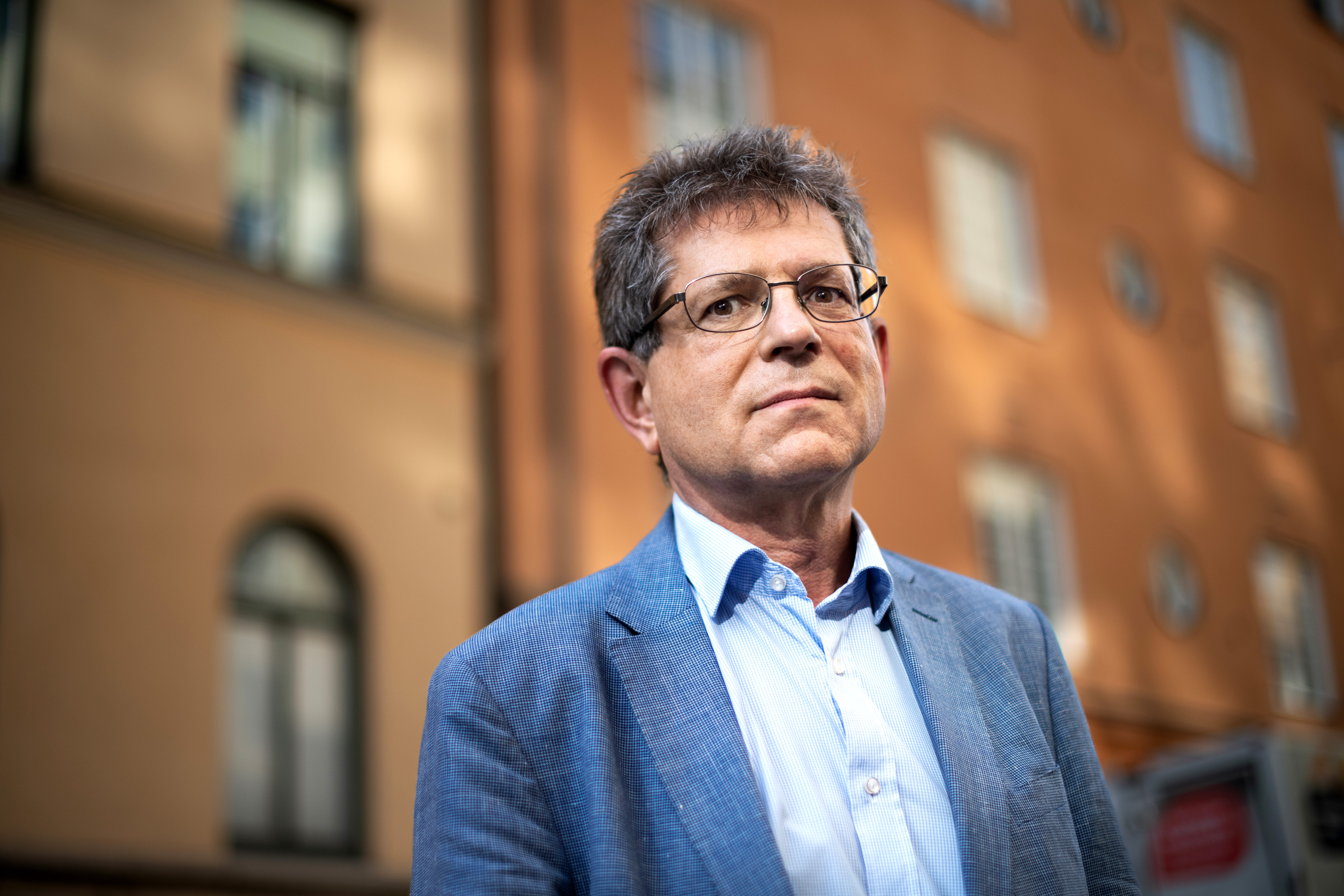 En person med glasögon och en blå kostym står utomhus utomhus en byggnad med orange väggar.