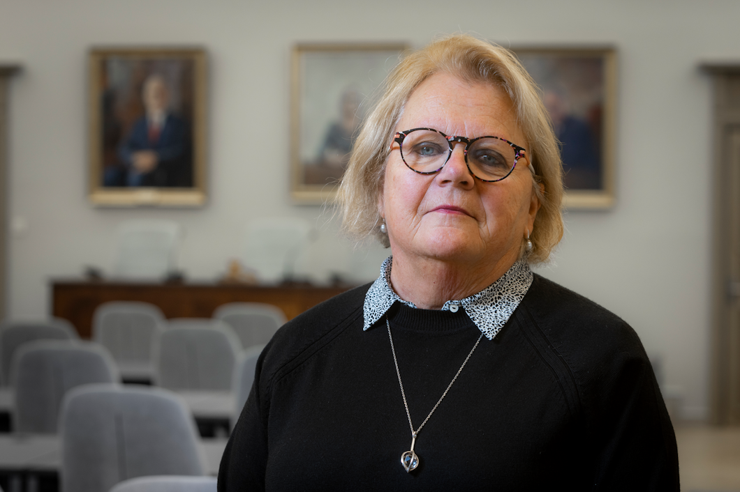 Marianne Henriksson med glasögon står i ett rum med grå stolar och inramade porträtt på väggarna.