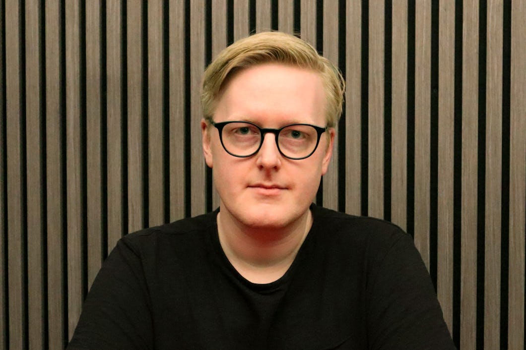 Stödassistenten Niklas Granath med kort blont hår och glasögon sitter framför en bakgrund med vertikala ränder, iförd en svart skjorta.
